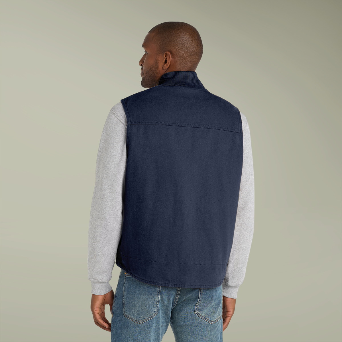 Man wearing a navy vest over a light gray long-sleeve shirt and blue jeans on a beige background