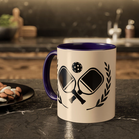 pickleball mug A ceramic mug with a cream exterior and dark purple interior, featuring a black graphic of crossed paddles, a ball, and laurel leaves. The mug is set on a dark marble countertop in a blurred kitchen environment.