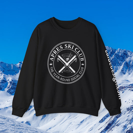 Apres Ski sweater A black crewneck sweatshirt with a white circular graphic reading 'APRES SKI CLUB' and crossed skis on the chest, plus white text down the sleeve, shown against a snowy mountain and blue sky background.