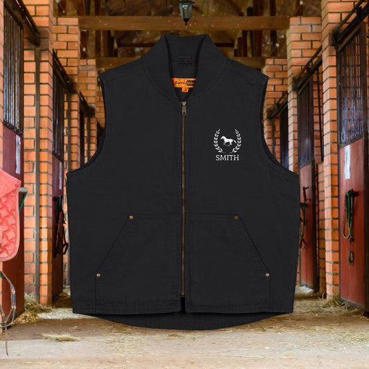 Equestrian vest A black sleeveless zip-front vest with a stand collar, front pockets, and a white embroidered horse logo with the word 'SMITH' on the left chest, shown hanging in a brick stable aisle.
