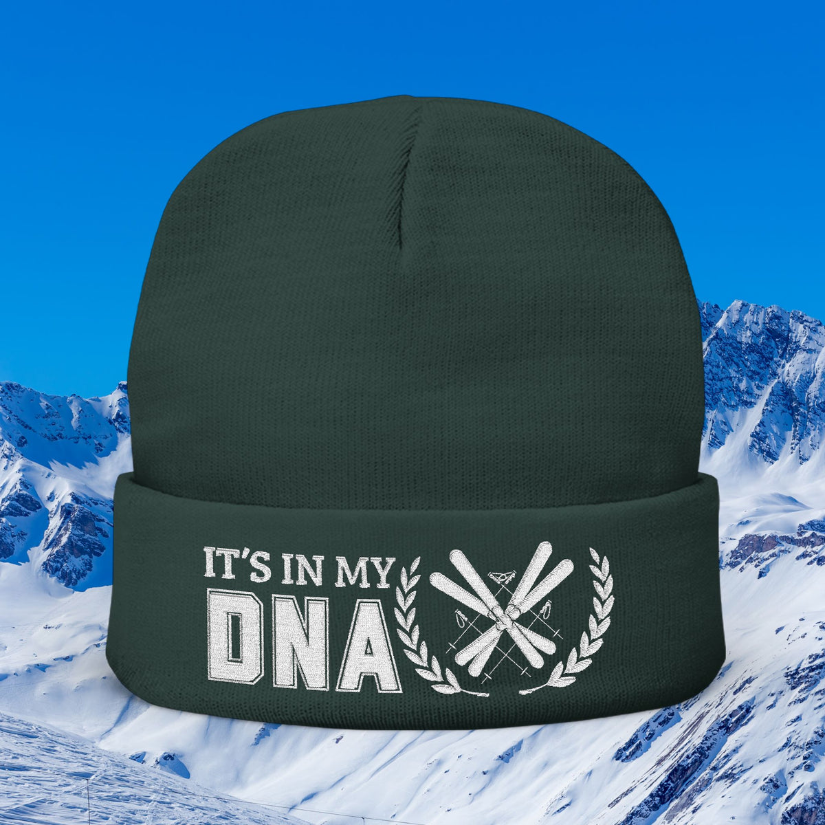 Ski Beanie A dark forest green knit beanie with a white graphic on the cuff that reads 'IT'S IN MY DNA' alongside crossed skis and a laurel wreath, set against a snowy mountain and blue sky background.