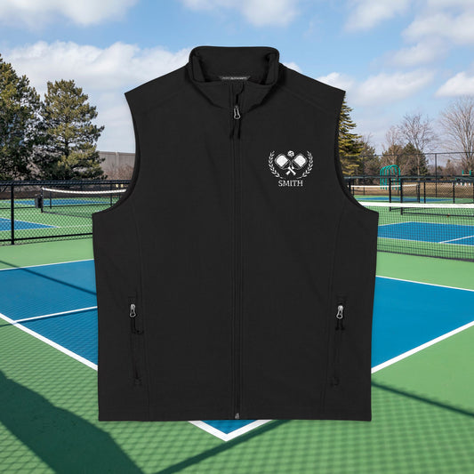 personalized pickleball gift A black sleeveless zip-front vest with a stand collar, shown centered over a tennis court background. The vest has a white chest graphic with crossed tennis rackets, laurel branches, and the name "SMITH" in a circular badge.