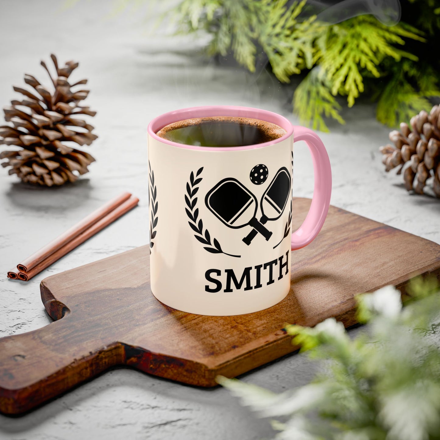 pickleball mug A personalized ceramic mug with a pink handle and rim, featuring a black ping-pong graphic and the name 'SMITH' printed on a cream body. The mug sits on a wooden cutting board surrounded by pinecones, cinnamon sticks, and green foliage on a gray stone surface.