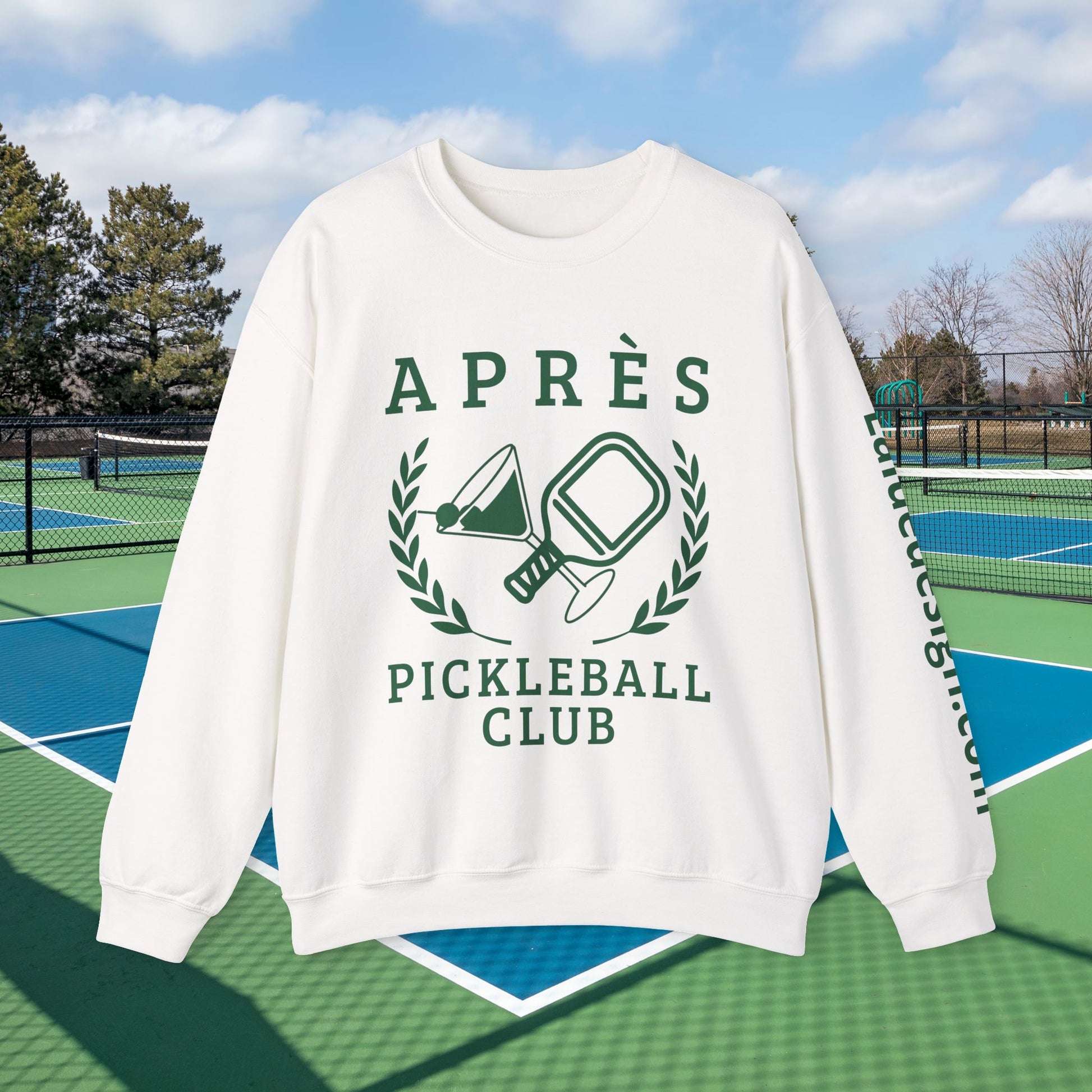 personalized padel gift An off-white crewneck sweatshirt with a dark green graphic print reading 'APRÈS PICKLEBALL CLUB' with pickleball rackets and a laurel wreath, shown against a background of outdoor pickleball courts under a blue sky.