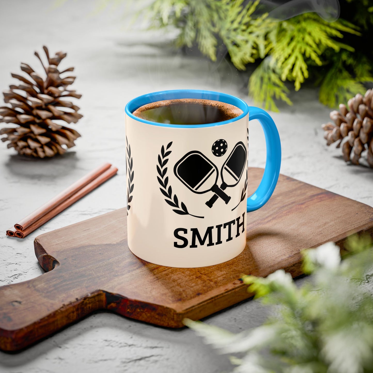 pickleball mug A personalized ceramic mug with a blue handle and rim sits on a wooden cutting board. The mug is cream-colored with a black ping-pong graphic and the name 'SMITH' printed on it.