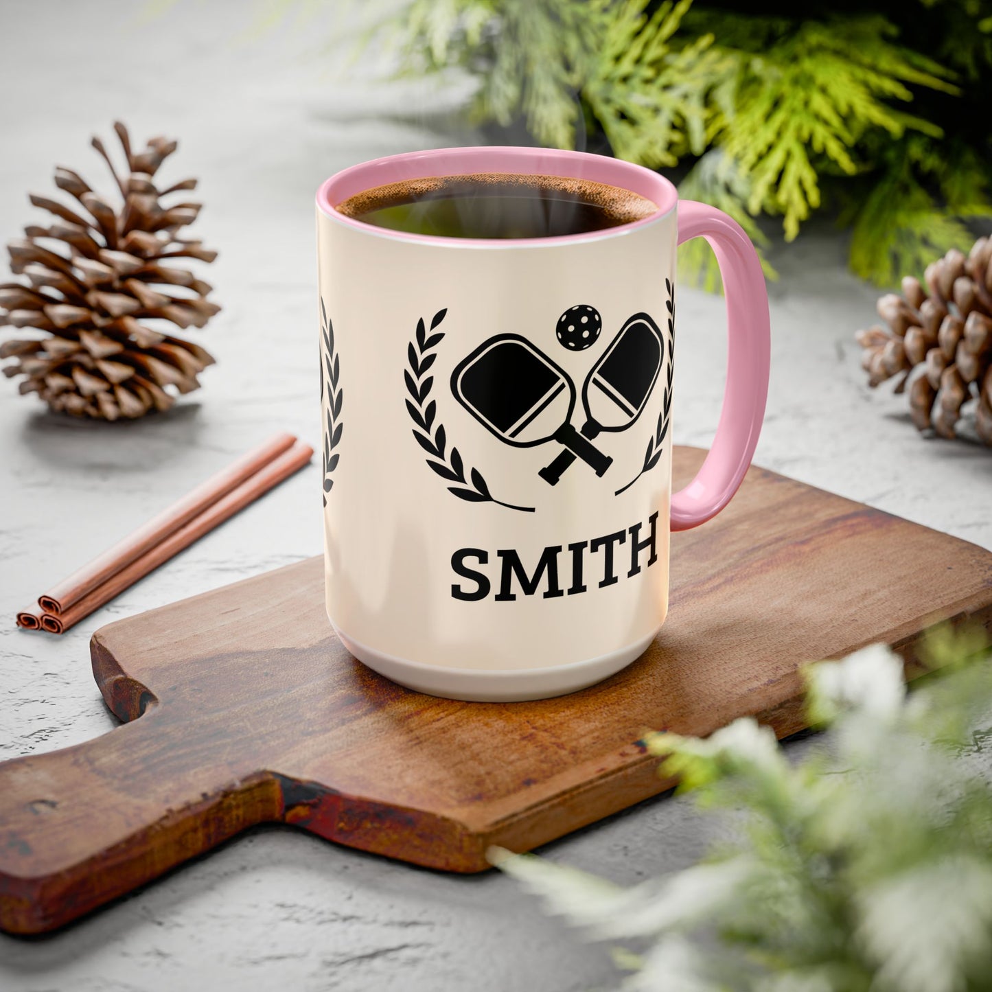 pickleball mug A personalized cream-colored ceramic mug with a pink handle and rim, filled with coffee and placed on a wooden cutting board. The mug features a black graphic of ping-pong paddles and the name 'SMITH' printed below.