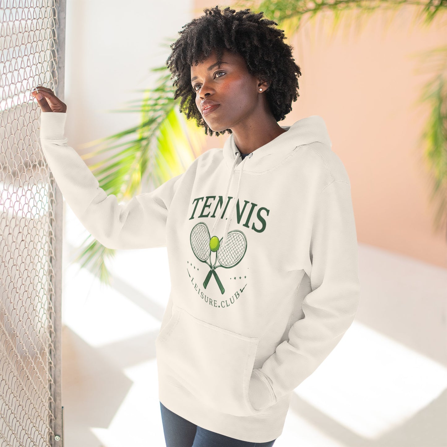 Person wearing a white hoodie with 'Tennis' and racket design, standing against a light background with plants.