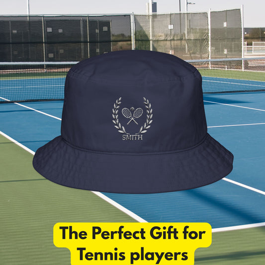 Tennis bucket hat A navy blue bucket hat with a white embroidered laurel wreath, crossed tennis rackets, and the name 'SMITH' on the front, shown against a tennis court background with a yellow promotional text banner at the bottom.