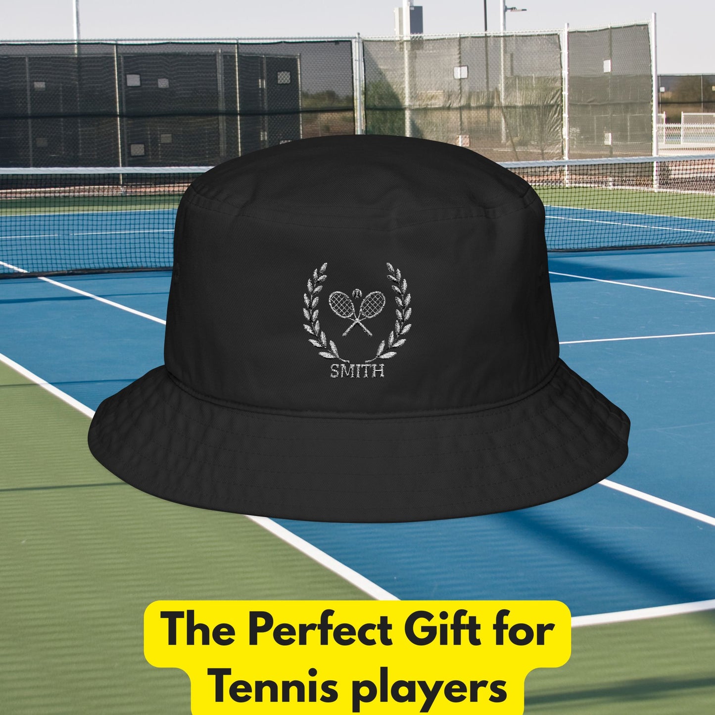 Tennis bucket hat A black bucket hat with a white laurel wreath and crossed tennis rackets graphic, featuring the name 'SMITH' below. The hat is superimposed over a tennis court background with a yellow promotional text banner at the bottom.