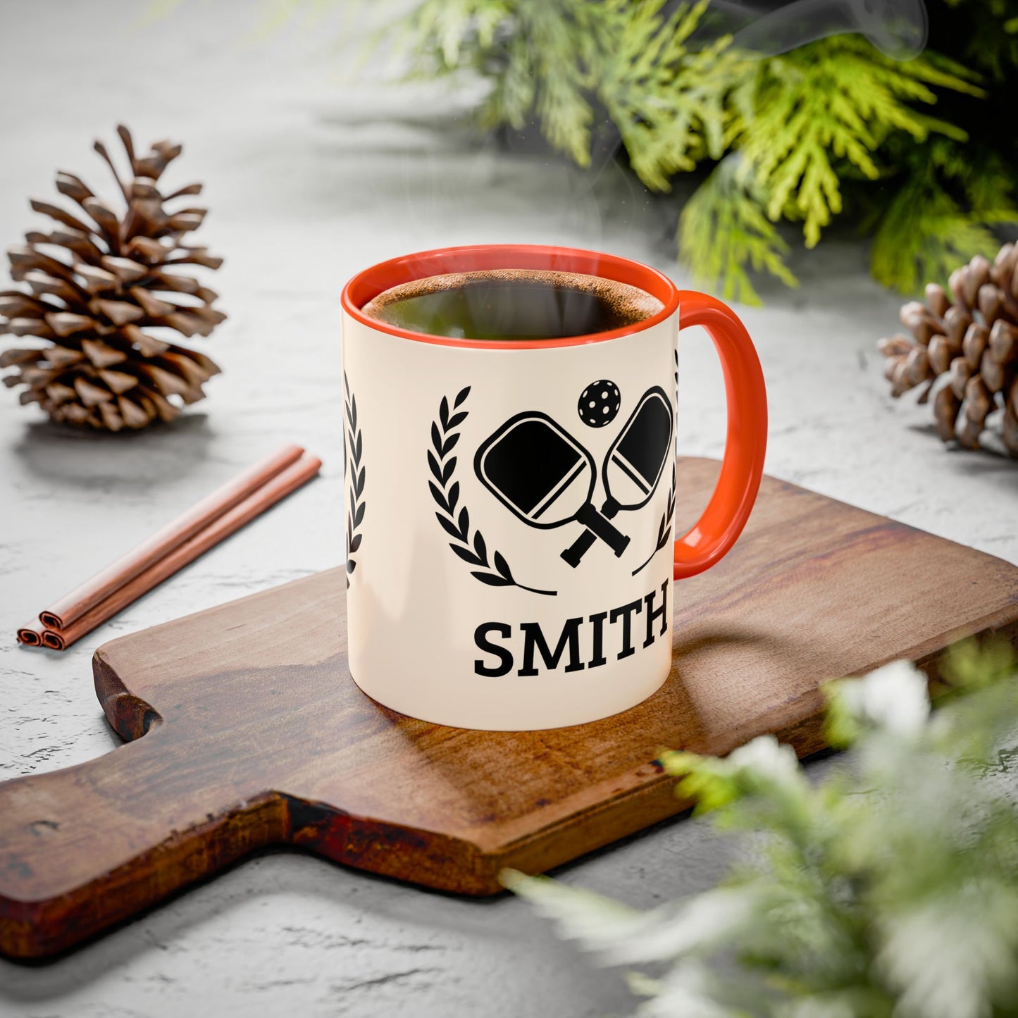 pickleball mug A personalized ceramic mug with an orange handle and rim, featuring a black ping-pong graphic and the name 'SMITH' printed on a wooden cutting board. The scene is styled with pinecones, cinnamon sticks, and green foliage on a gray stone surface.