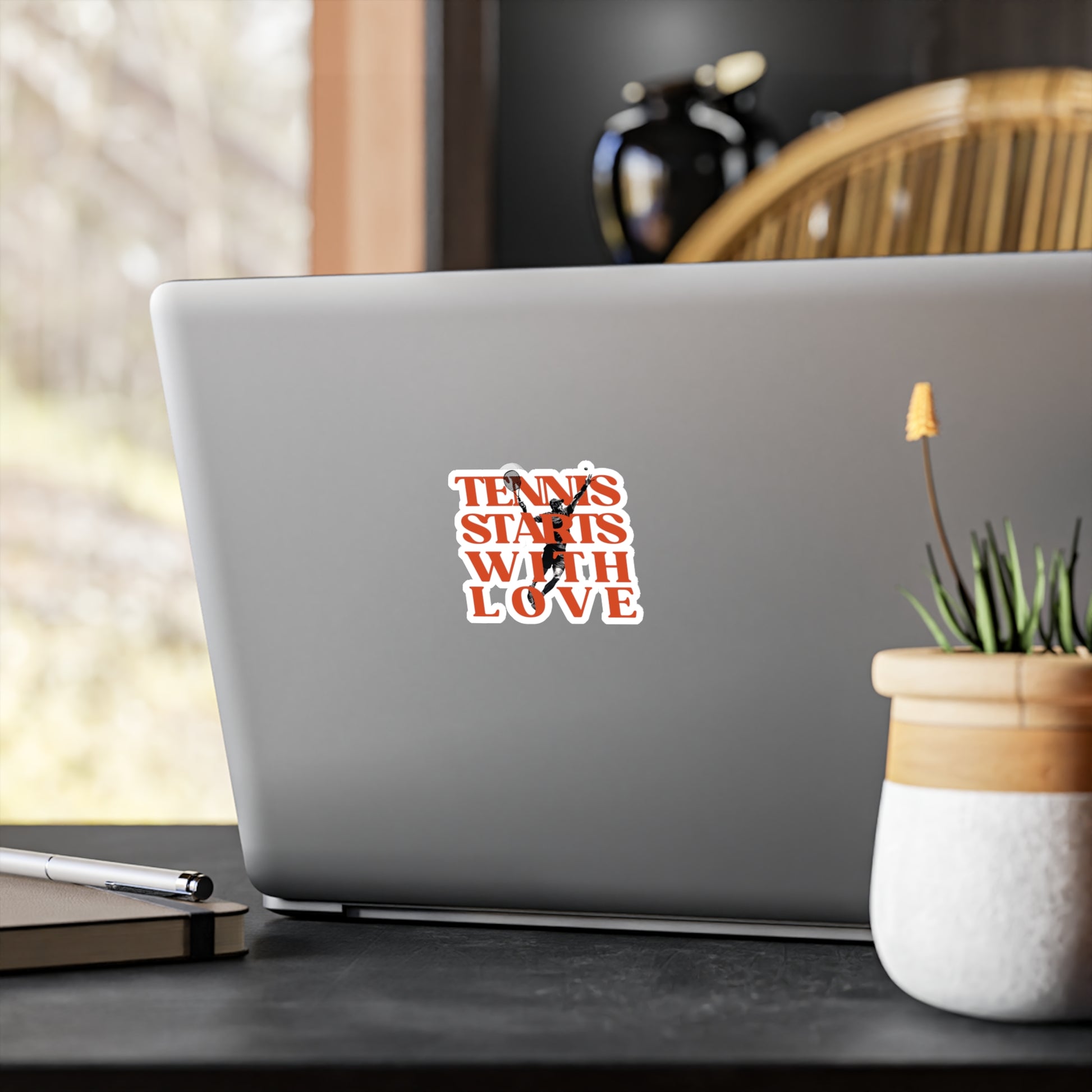 Laptop with a 'Tennis Starts with Love' sticker on a desk with a plant and pen.