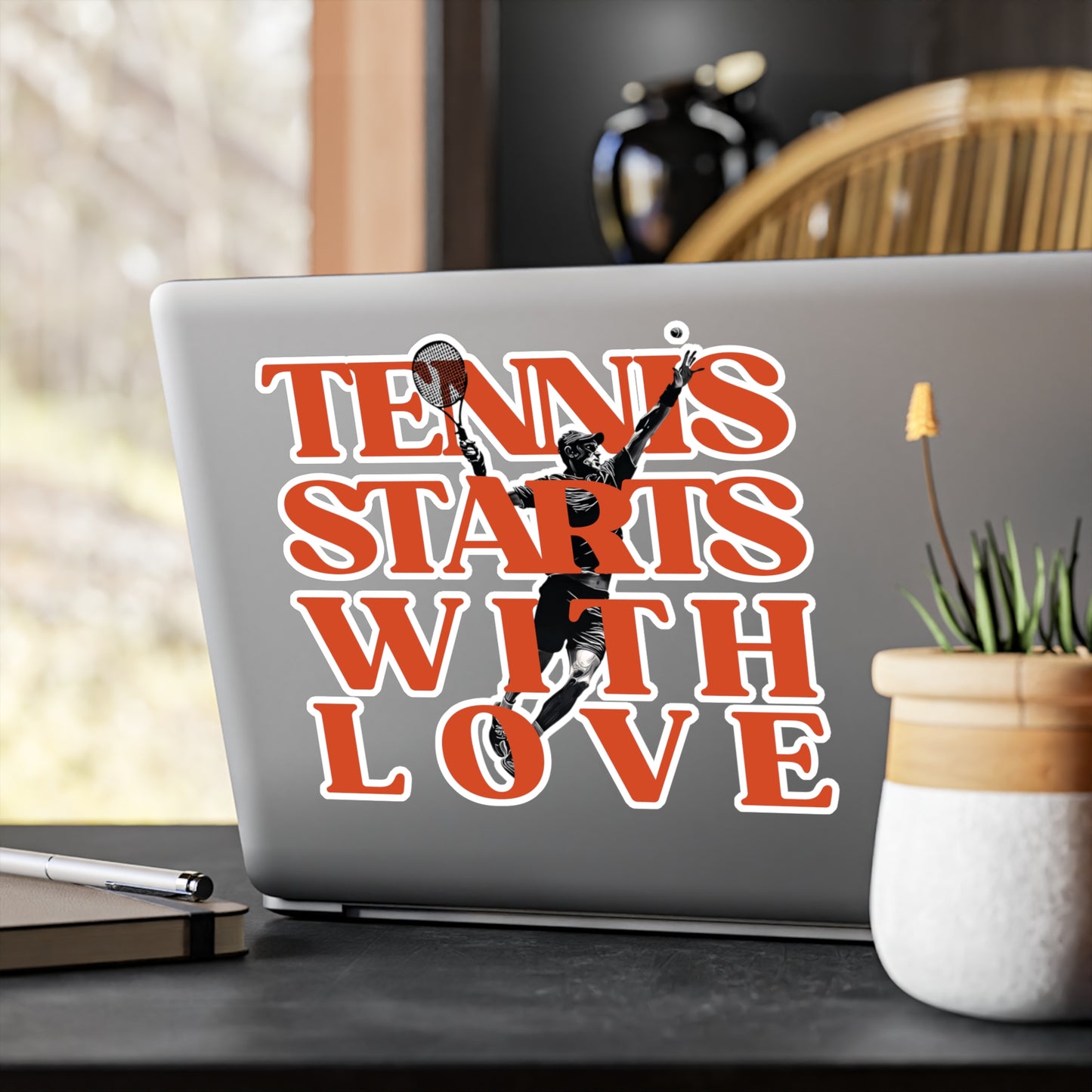 Laptop with 'Tennis Starts with Love' sticker on a desk with a plant and pen.
