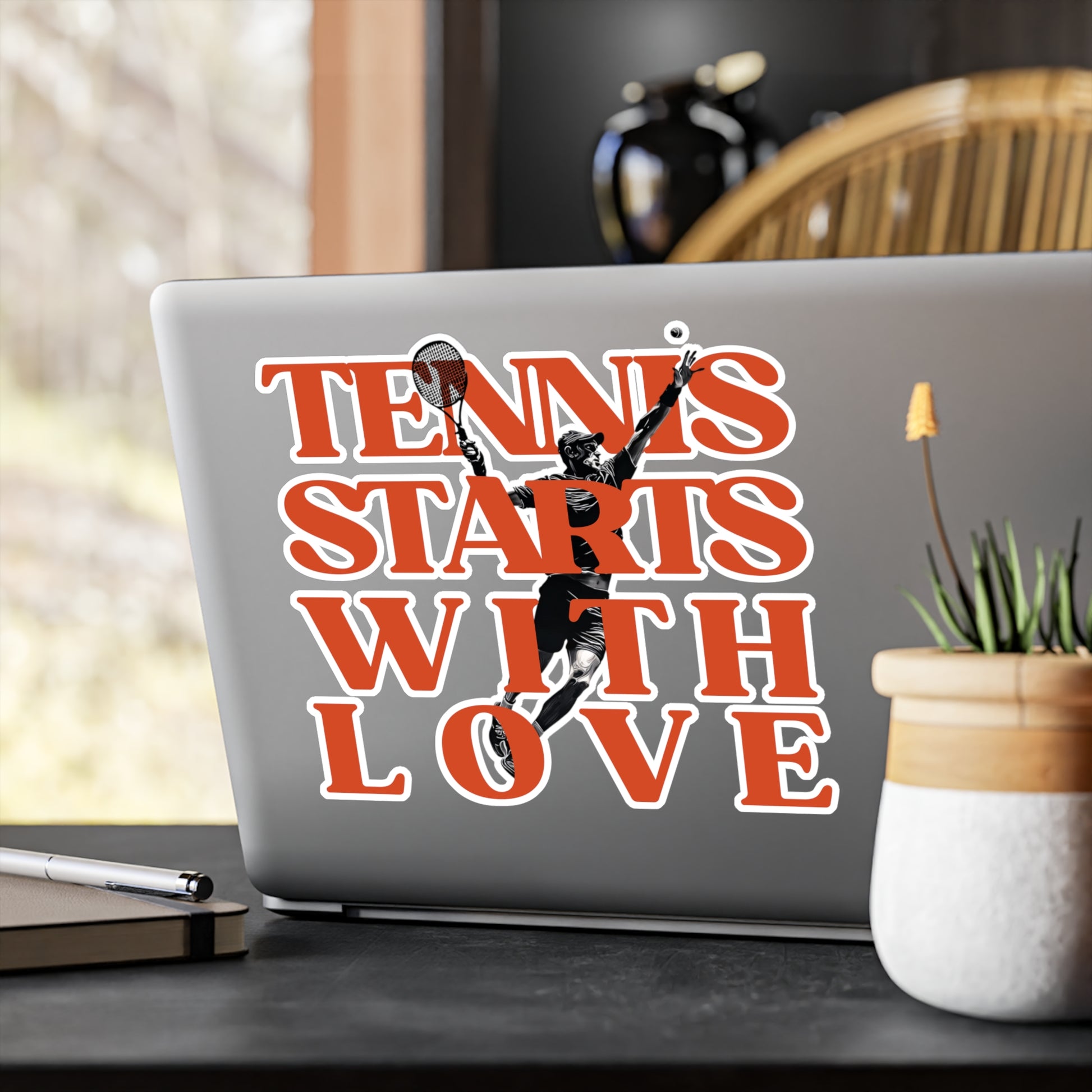 Laptop with 'Tennis Starts with Love' sticker on a desk with a plant and pen.