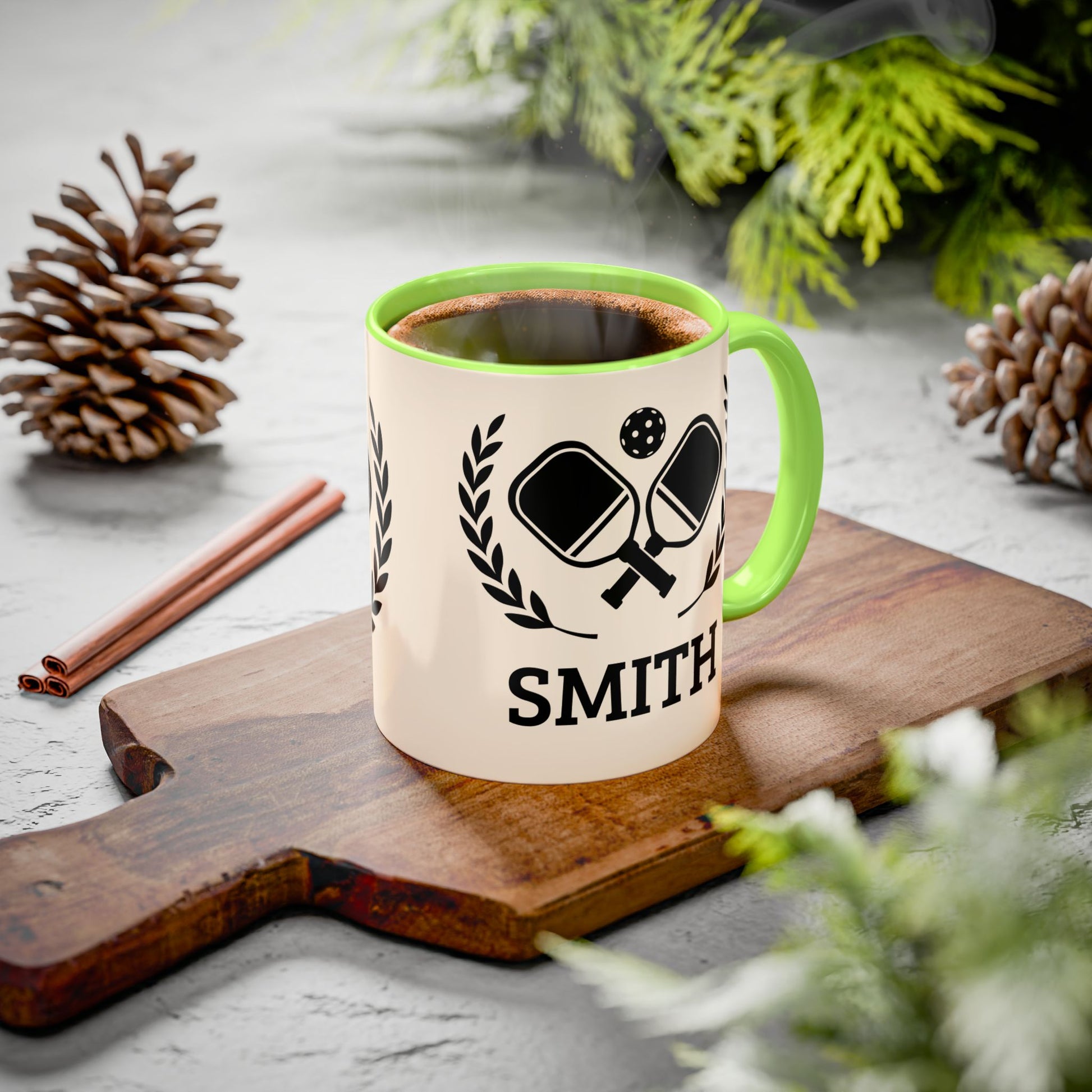 pickleball mug A personalized ceramic mug with a green handle and interior, featuring a black ping-pong graphic and the name 'SMITH' printed on a wooden cutting board. The scene is styled with pinecones, cinnamon sticks, and green foliage on a gray surface.