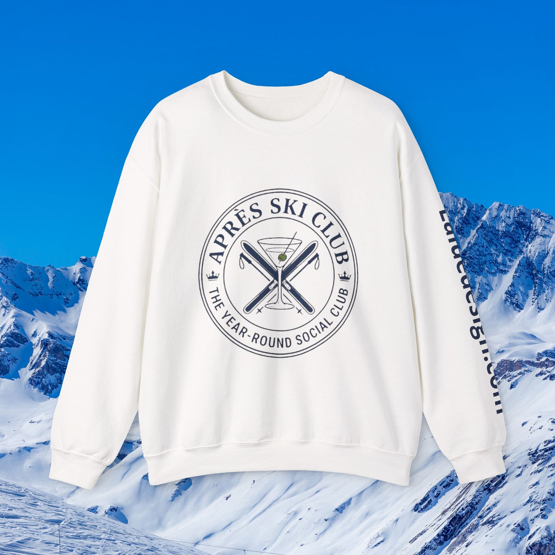 Après Ski Sweatshirt An off-white crewneck sweatshirt with a circular graphic on the chest reading 'APRÈS SKI CLUB THE YEAR-ROUND SOCIAL CLUB' around crossed skis and a martini glass, shown against a snowy mountain background with a bright blue sky.