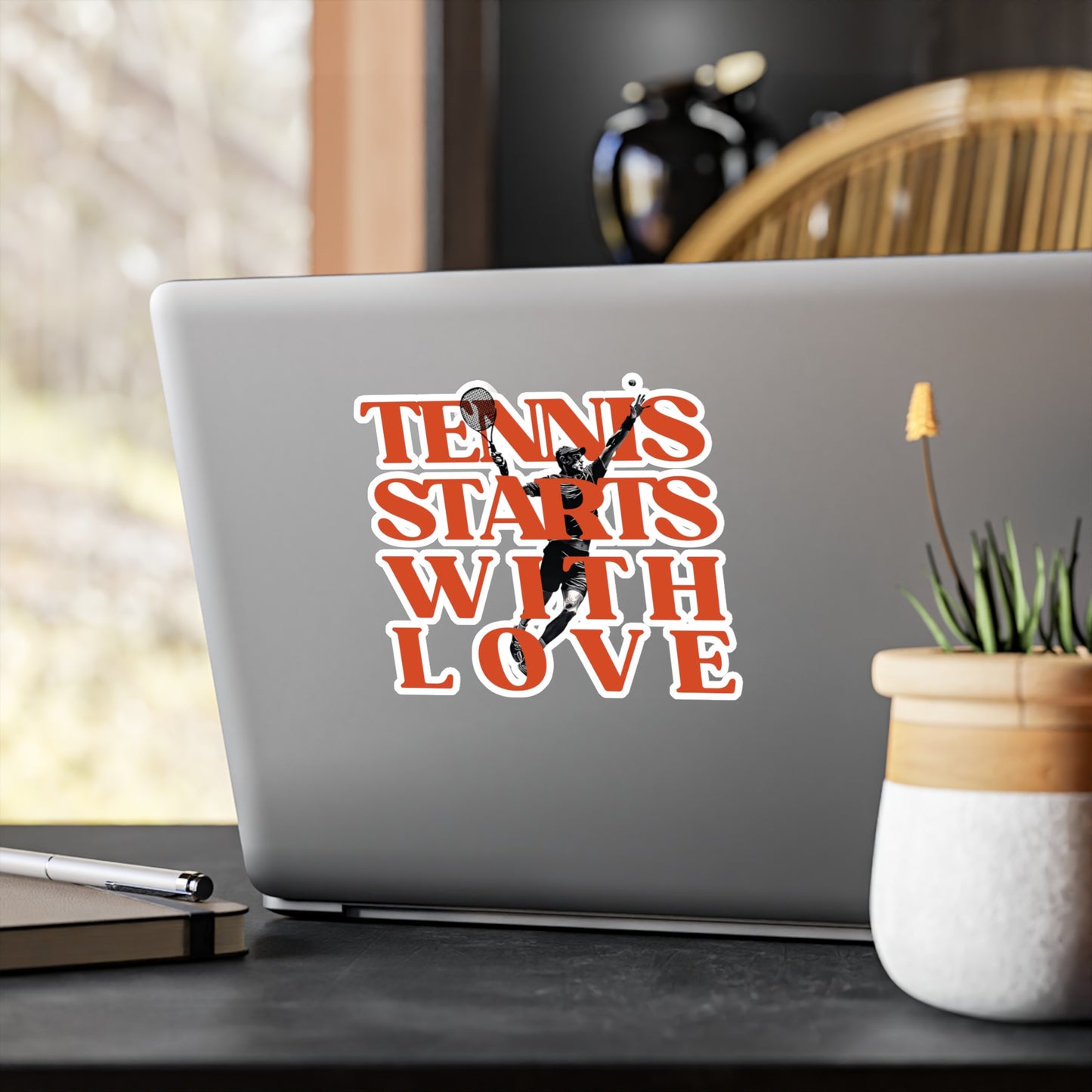 Laptop with a 'Tennis Starts with Love' sticker on a desk with a plant and pen.