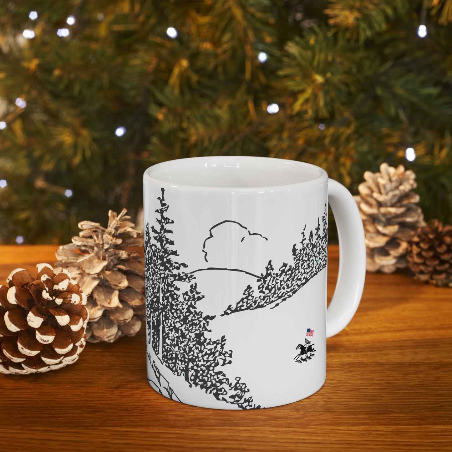 White mug with black landscape design on a wooden surface with pinecones and a Christmas tree in the background.