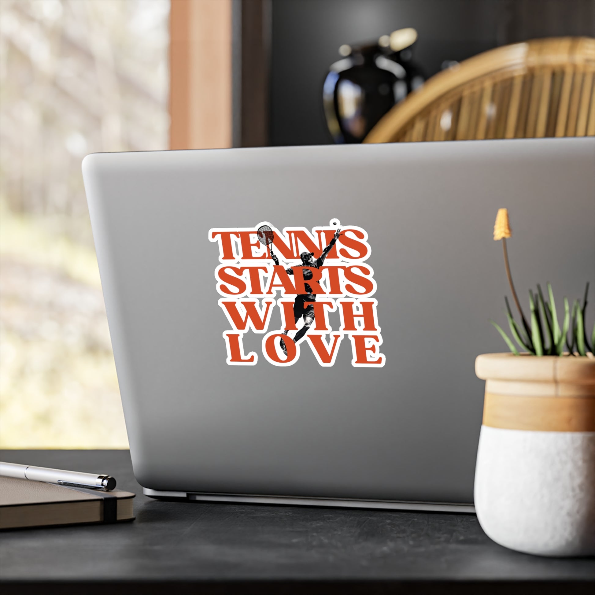 Laptop with 'Tennis Starts with Love' sticker on a desk with a plant and pen.