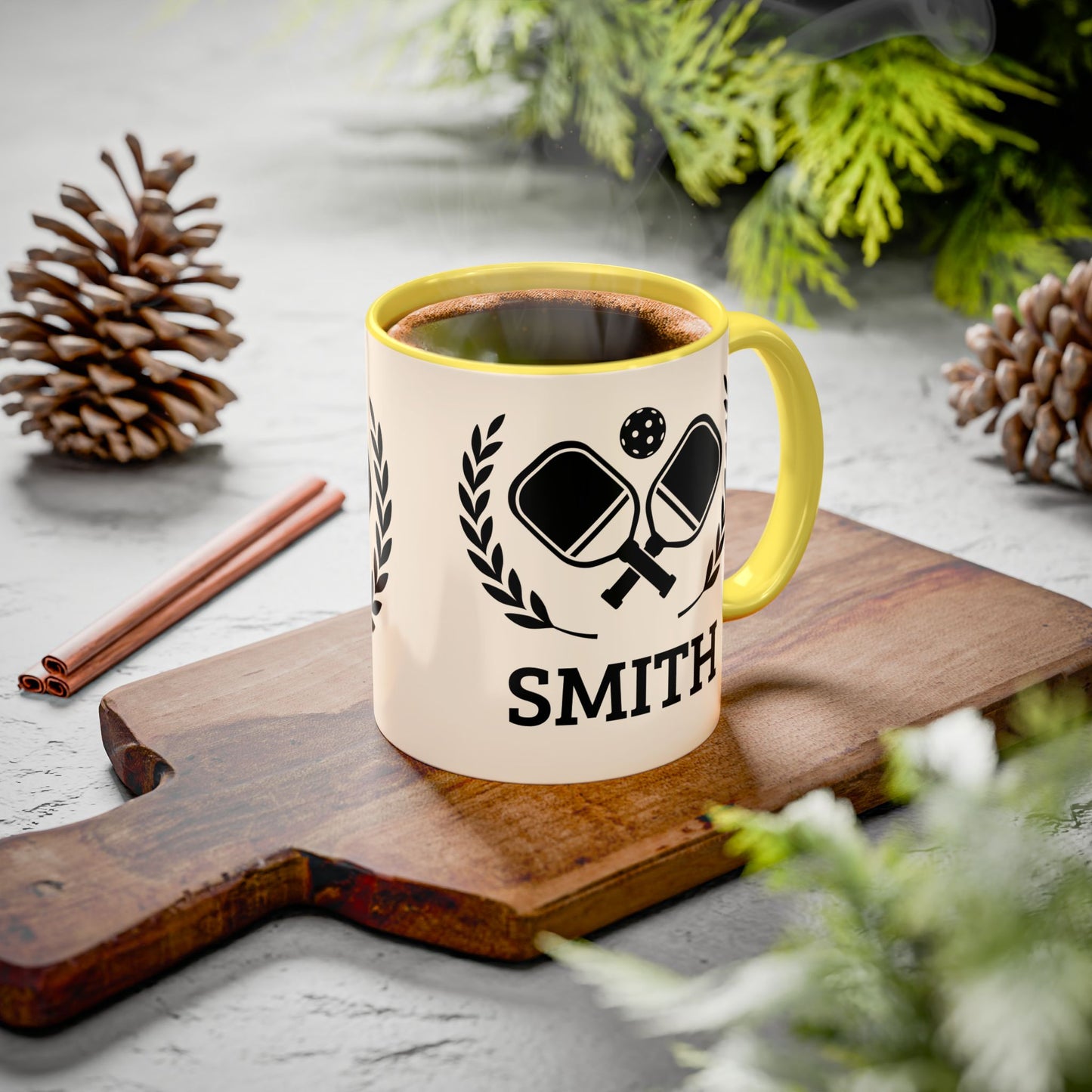 pickleball mug A personalized ceramic mug with a green handle and interior, featuring a black ping-pong graphic and the name 'SMITH' printed on a wooden cutting board. The scene is styled with pinecones, cinnamon sticks, and green foliage on a gray surface.