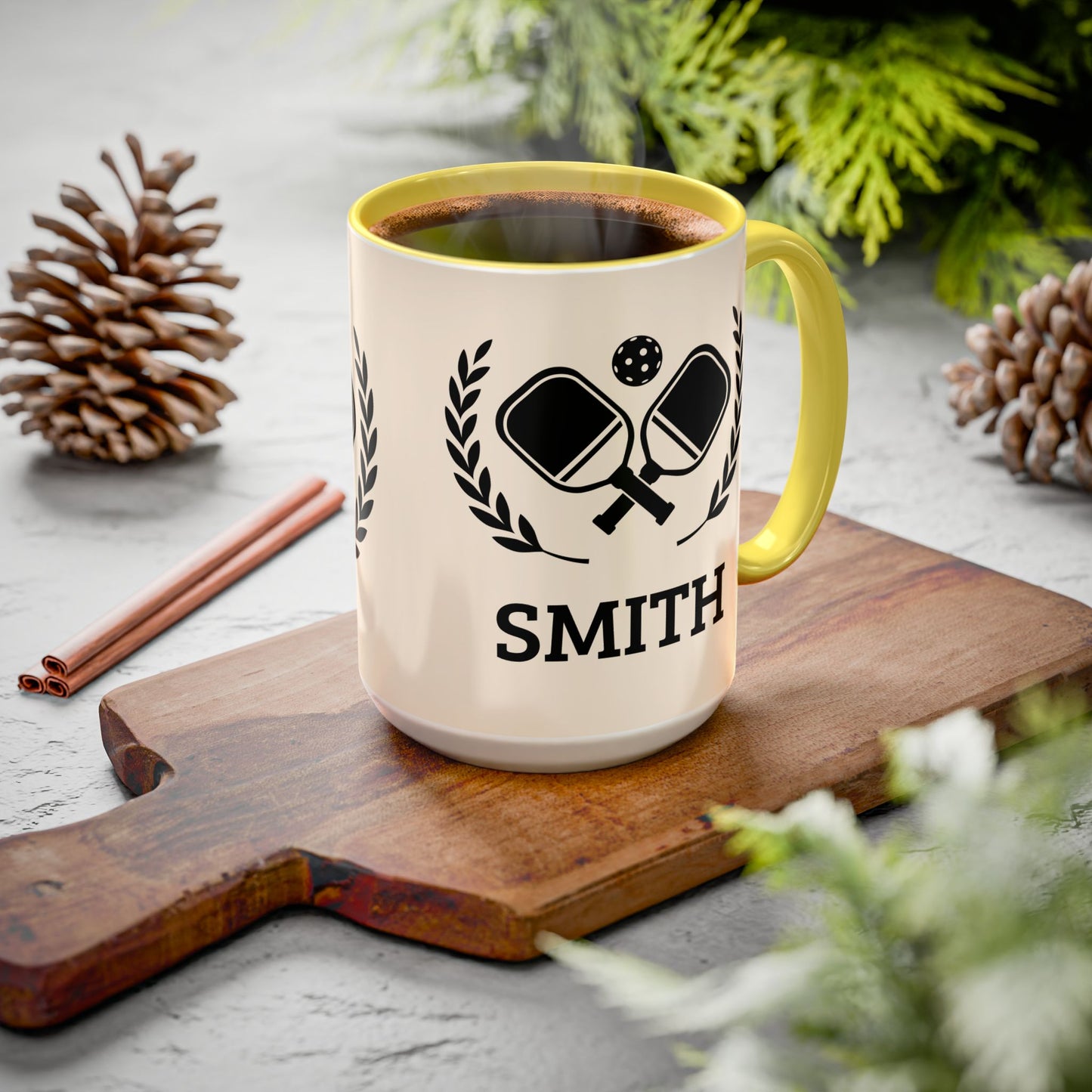 pickleball mug A personalized ceramic mug with a yellow handle and rim, filled with coffee, sitting on a wooden cutting board. The mug is cream-colored with a black graphic of ping-pong paddles and the name 'SMITH' printed below.