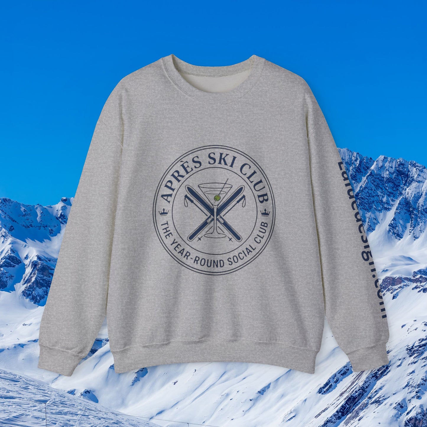 Après Ski Sweatshirt A light gray crewneck sweatshirt with a dark gray circular graphic on the chest reading 'APRÈS SKI CLUB THE YEAR-ROUND SOCIAL CLUB' around crossed skis and a martini glass, with 'Lalique Ski Club' printed down the sleeve. The sweatshirt is displayed against a digital background of snowy mountains and a blue sky.