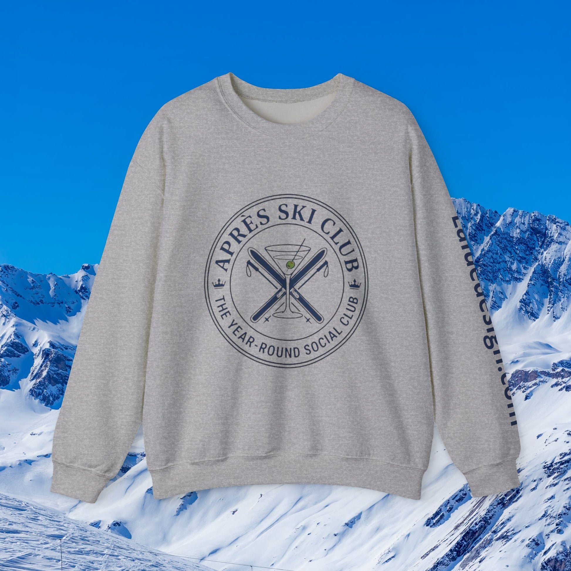 Après Ski Sweatshirt A light gray crewneck sweatshirt with a dark gray circular graphic on the chest reading 'APRÈS SKI CLUB THE YEAR-ROUND SOCIAL CLUB' around crossed skis and a martini glass, with 'Lalique Ski Club' printed down the sleeve. The sweatshirt is displayed against a digital background of snowy mountains and a blue sky.