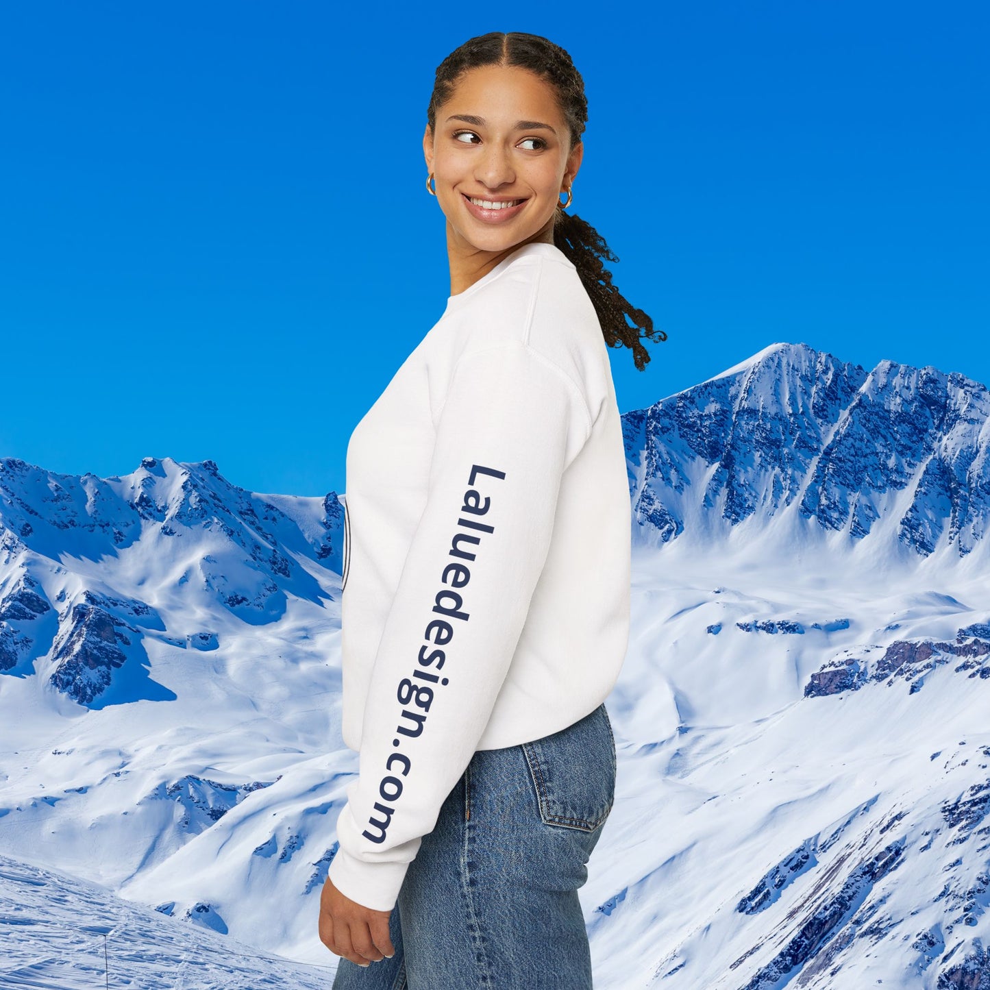 Après Ski Sweatshirt A woman wearing a white crewneck sweatshirt with 'Laluedesign.com' printed vertically in navy on the left sleeve, posed against a bright blue sky and snowy mountain backdrop.