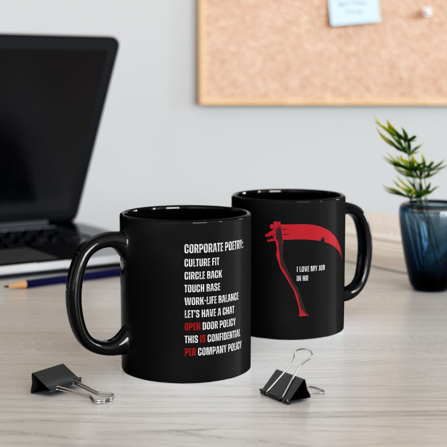Two black mugs with text on a desk with office items and a plant in the background.