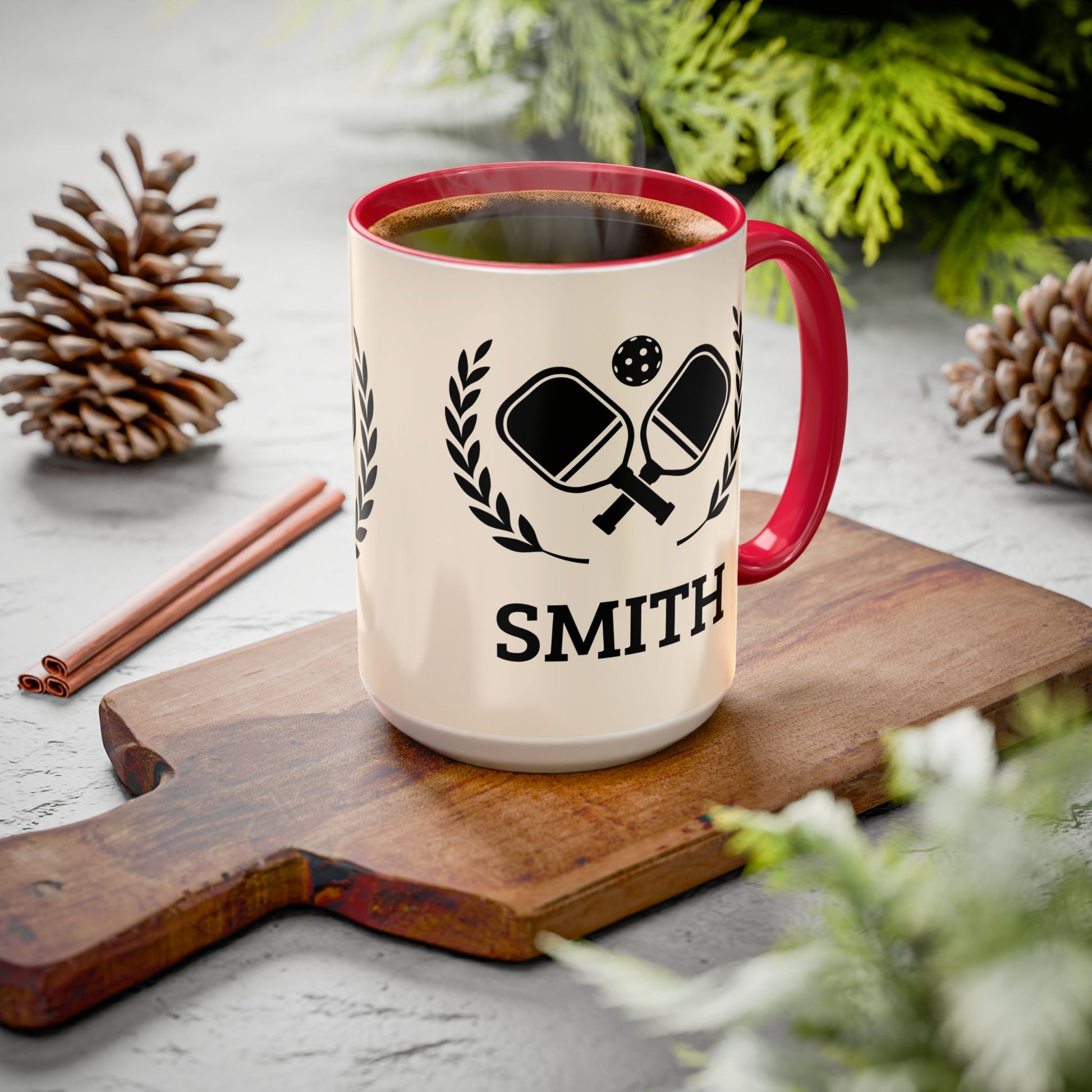 pickleball mug A personalized ceramic coffee mug with a red handle and rim, filled with coffee, sitting on a wooden cutting board. The mug is cream-colored with a black graphic of ping-pong paddles and the name 'SMITH' printed below.