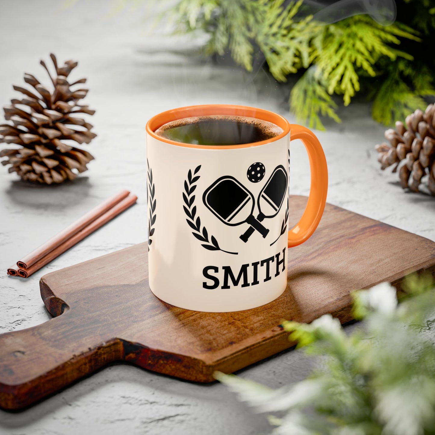 pickleball mug A personalized ceramic mug with an orange handle and rim, featuring a black ping-pong graphic and the name 'SMITH' printed on a wooden cutting board. The scene is styled with pinecones, cinnamon sticks, and green foliage on a gray stone surface.