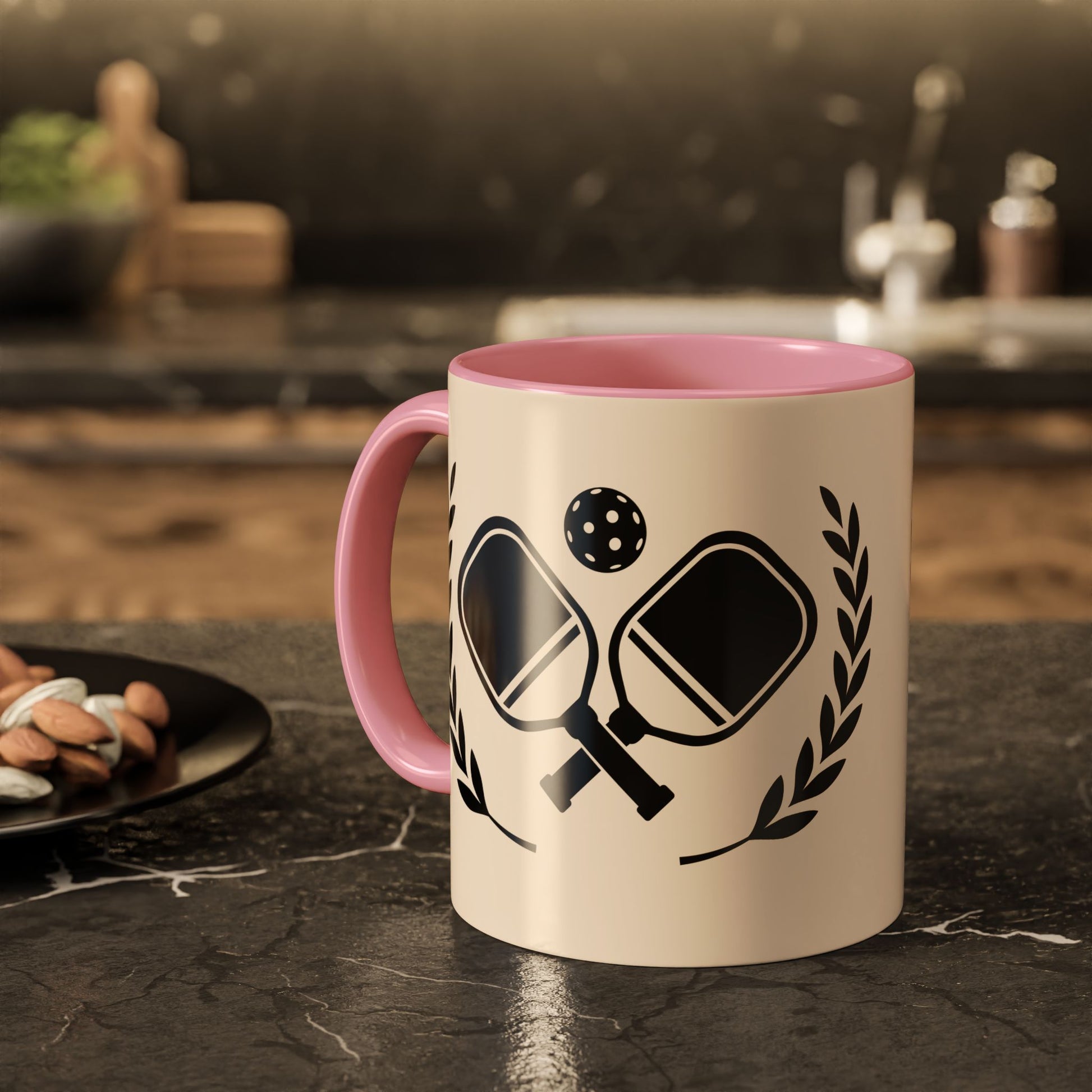 pickleball mug A ceramic mug with a pink interior and handle, featuring a black graphic of table tennis paddles, a ball, and laurel leaves on a cream-colored exterior, set on a dark kitchen countertop.