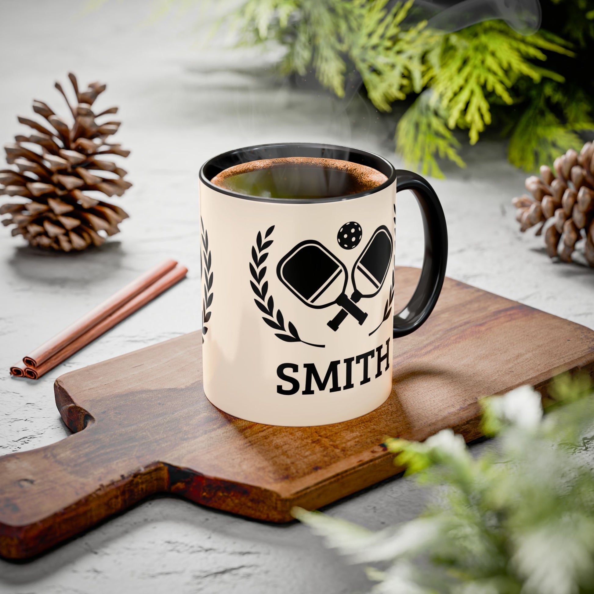 pickleball mug A cream-colored ceramic mug with a black handle and rim, featuring a black graphic of ping-pong paddles and the name 'SMITH' printed below. The mug is styled on a wooden cutting board with pinecones, cinnamon sticks, and green foliage on a gray surface.