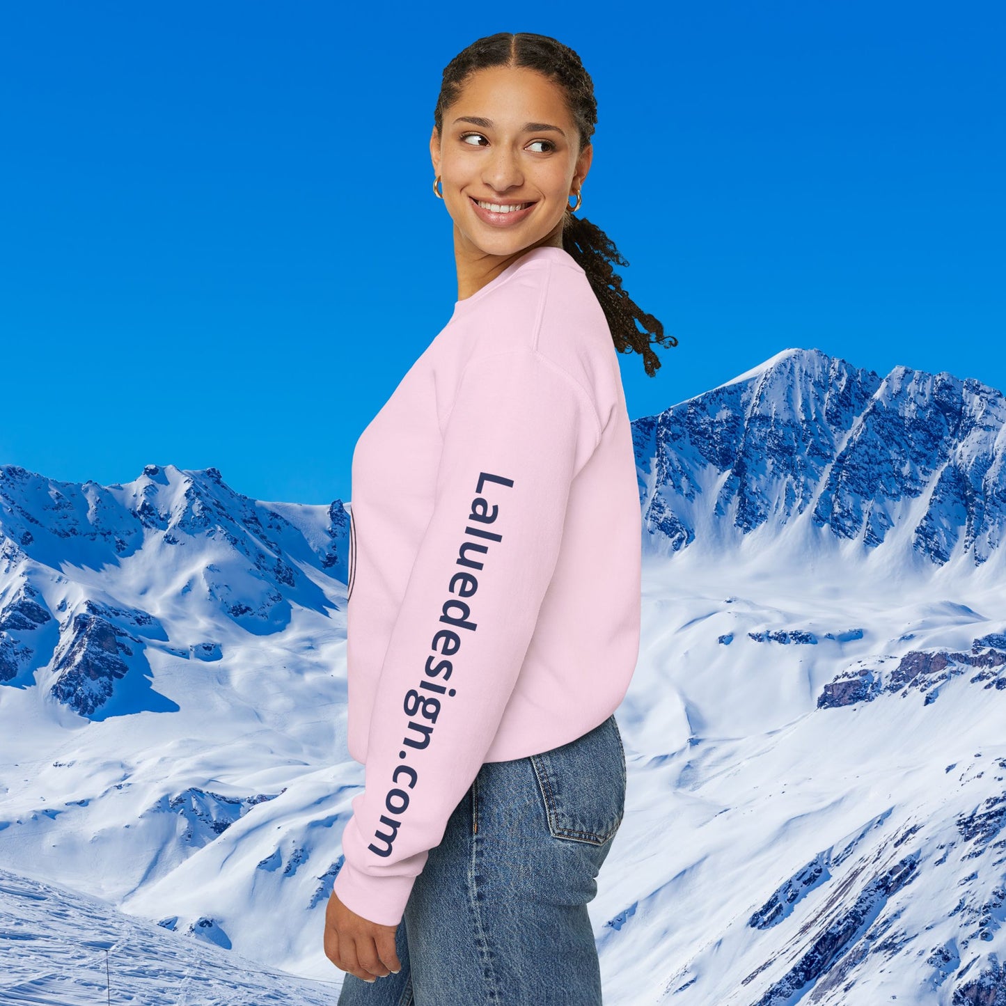 Après Ski Sweatshirt A woman wearing a light pink crewneck sweatshirt with 'Laliedesign.com' printed vertically in navy on the left sleeve, posed against a bright blue sky and snowy mountain backdrop.