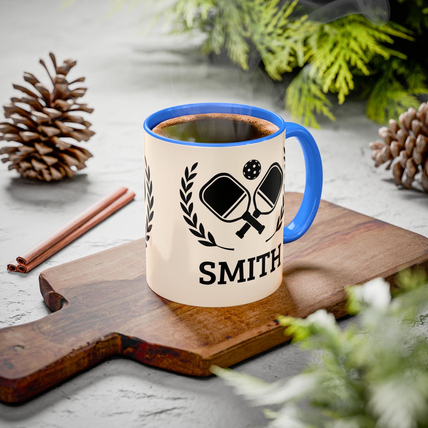 pickleball mug A ceramic mug with a blue handle and rim sits on a wooden cutting board, filled with a hot beverage and surrounded by pinecones, cinnamon sticks, and greenery on a light gray surface.