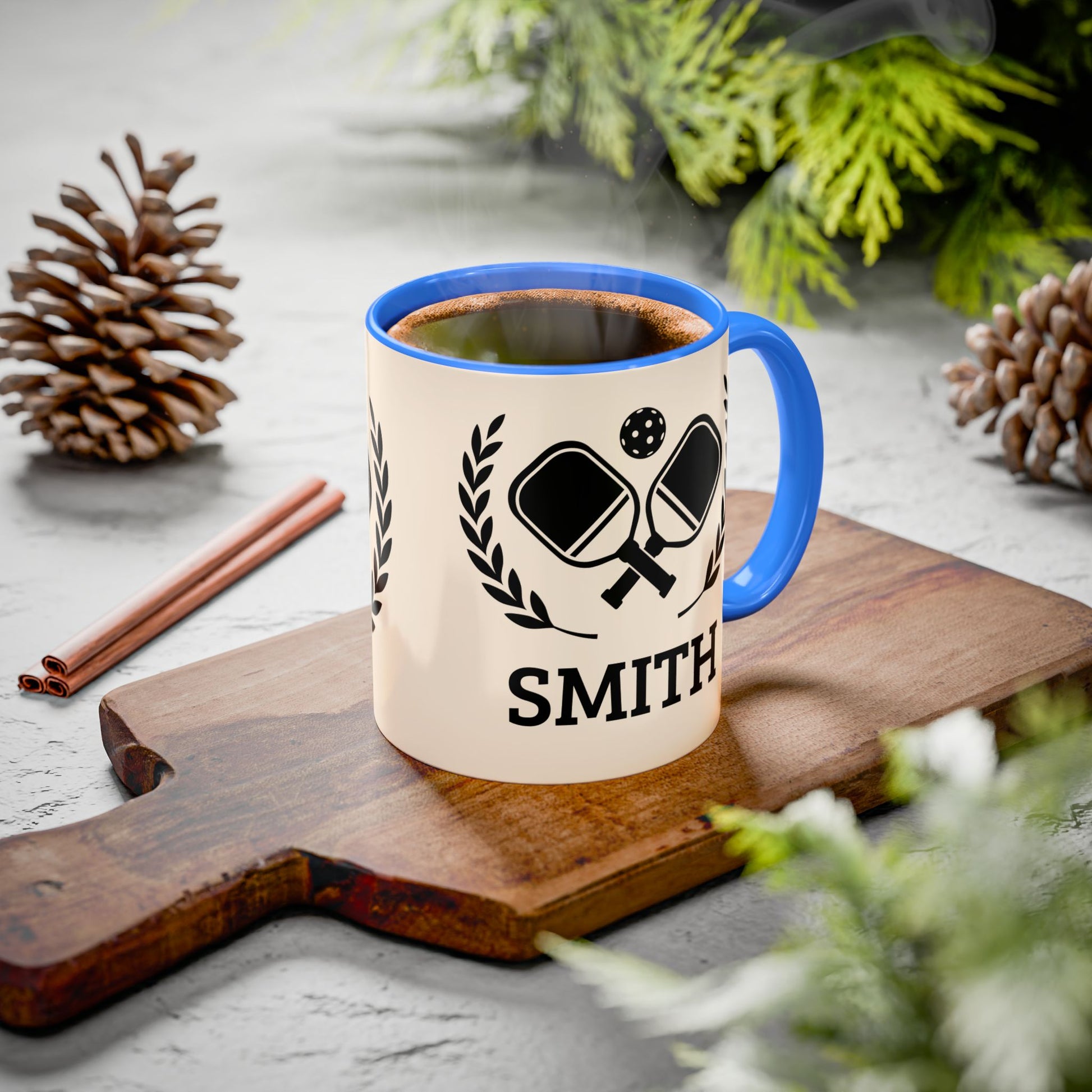 pickleball mug A ceramic mug with a blue handle and rim sits on a wooden cutting board, filled with a hot beverage and surrounded by pinecones, cinnamon sticks, and greenery on a light gray surface.