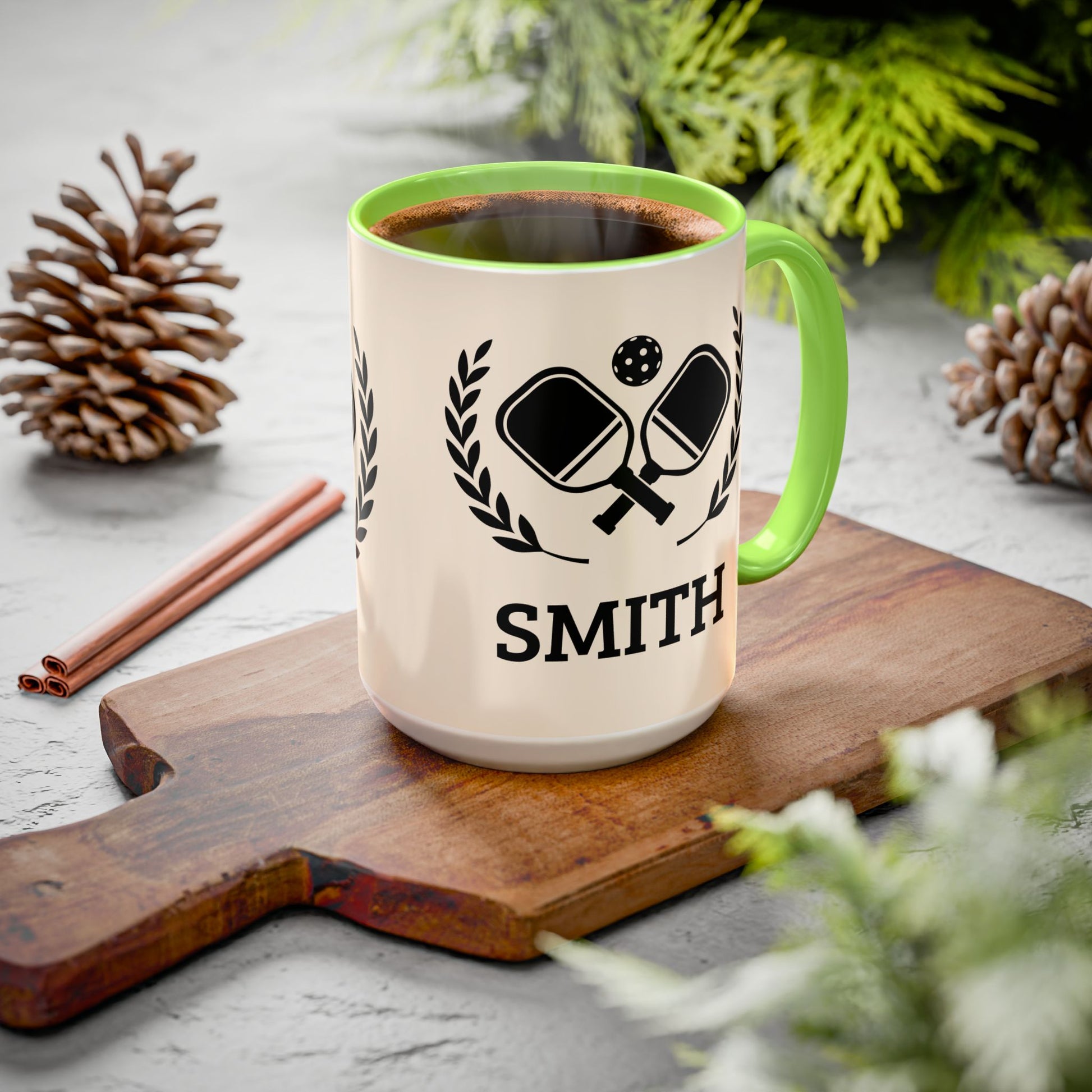 pickleball mug A ceramic mug with a cream-colored exterior, a bright green handle and interior rim, and a black printed graphic of crossed ping-pong paddles with the name 'SMITH' below. The mug is filled with a hot beverage and styled on a wooden cutting board with pinecones and greenery in the background.