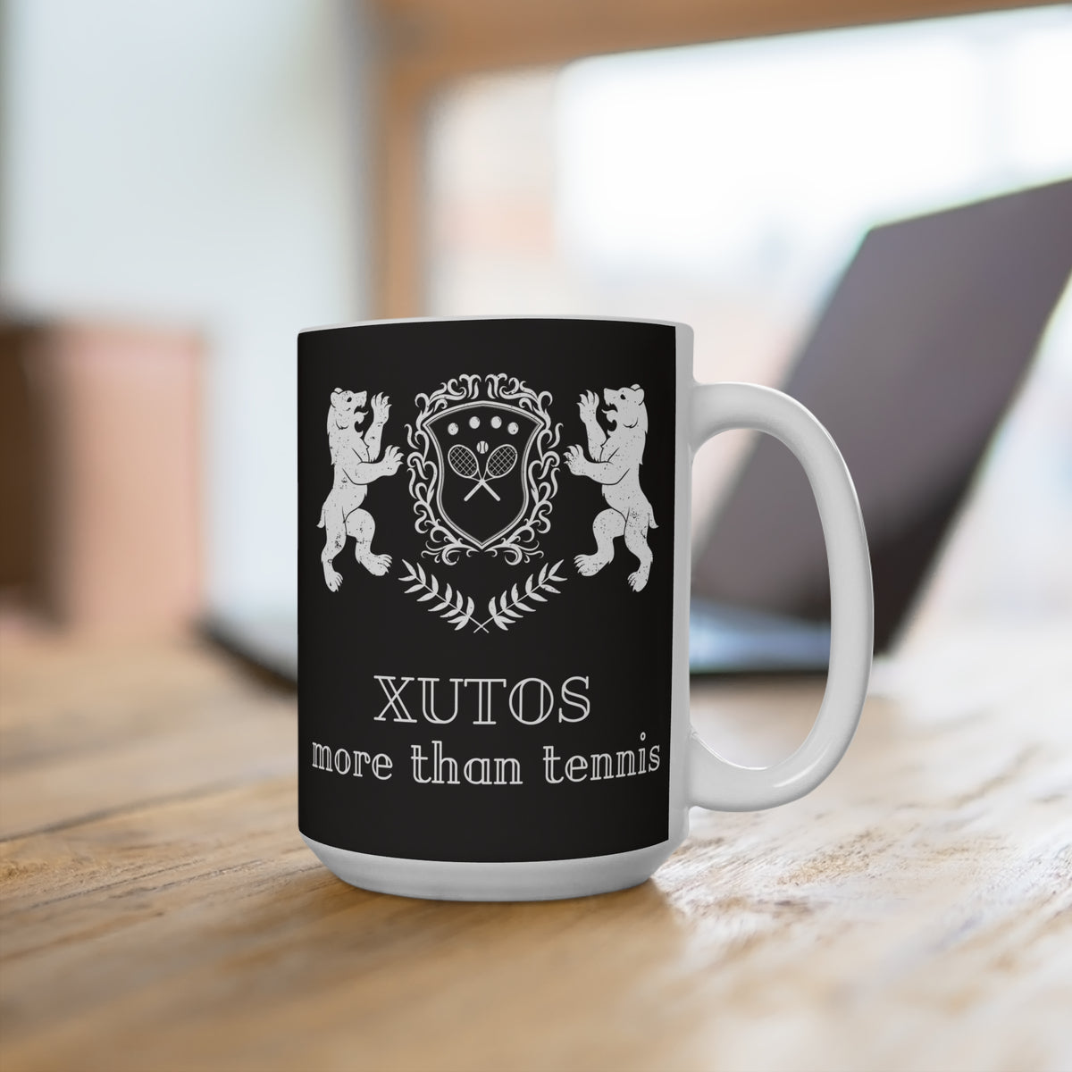 A white ceramic mug with a black printed wrap design featuring a tennis-themed crest and the text 'XUTOS more than tennis', shown on a wooden table with a blurred laptop in the background.