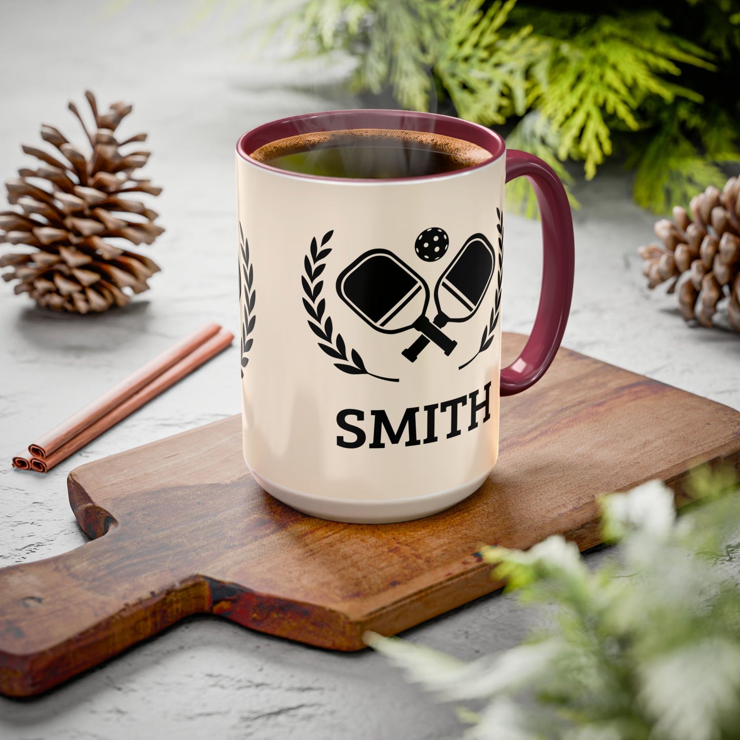 pickleball mug A cream-colored ceramic mug with a dark red handle and rim, featuring a black graphic of crossed ping-pong paddles and the name 'SMITH' printed below. The mug is filled with coffee and sits on a wooden cutting board surrounded by pinecones and greenery.
