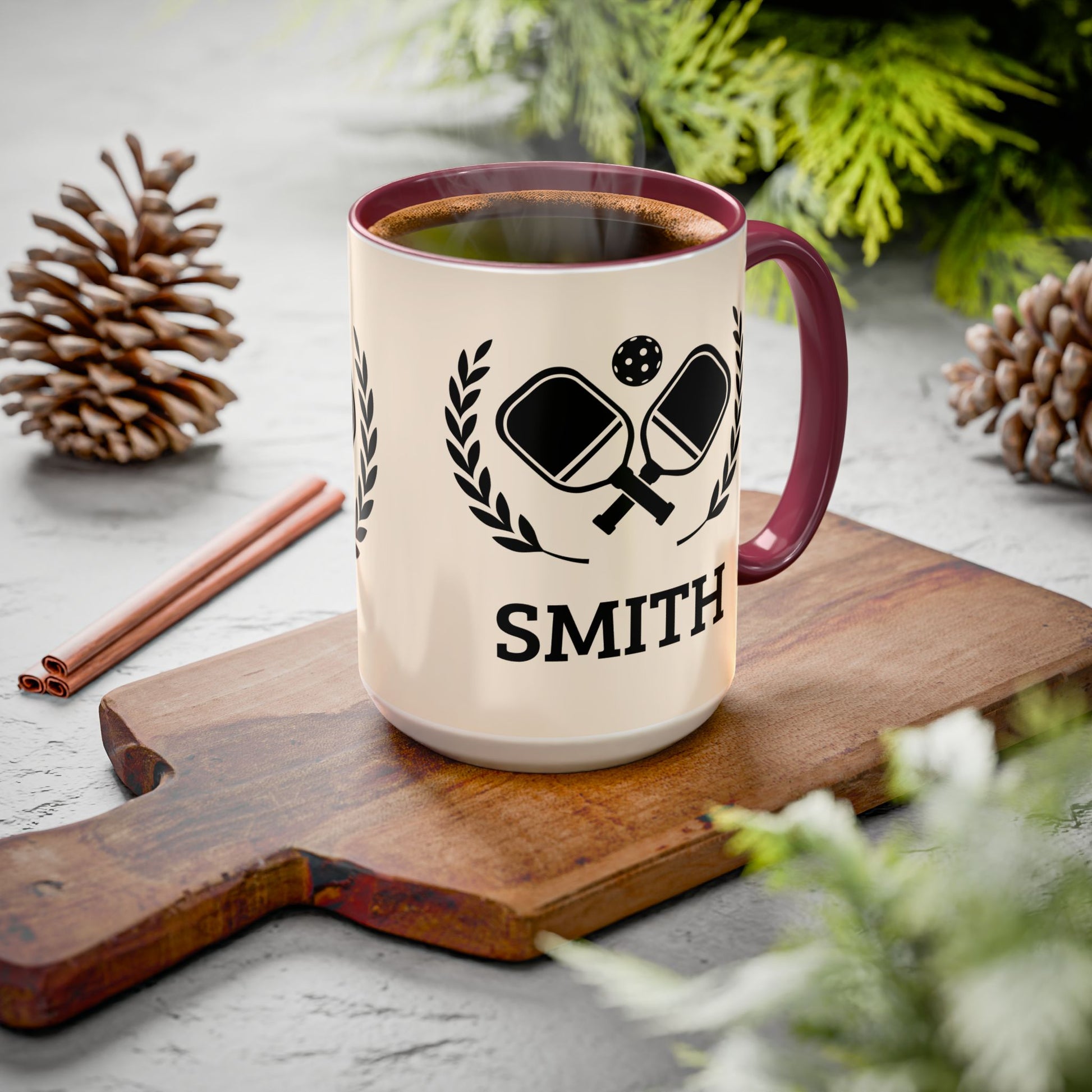 pickleball mug A cream-colored ceramic mug with a dark red handle and rim, featuring a black graphic of crossed ping-pong paddles and the name 'SMITH' printed below. The mug is filled with coffee and sits on a wooden cutting board surrounded by pinecones and greenery.