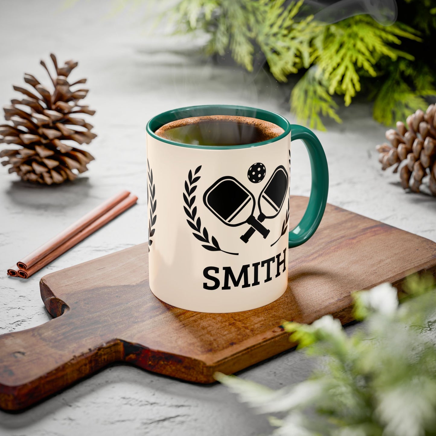 pickleball mug A ceramic mug with a green handle and rim sits on a wooden cutting board, filled with a hot beverage and surrounded by pinecones, cinnamon sticks, and greenery on a light gray surface.