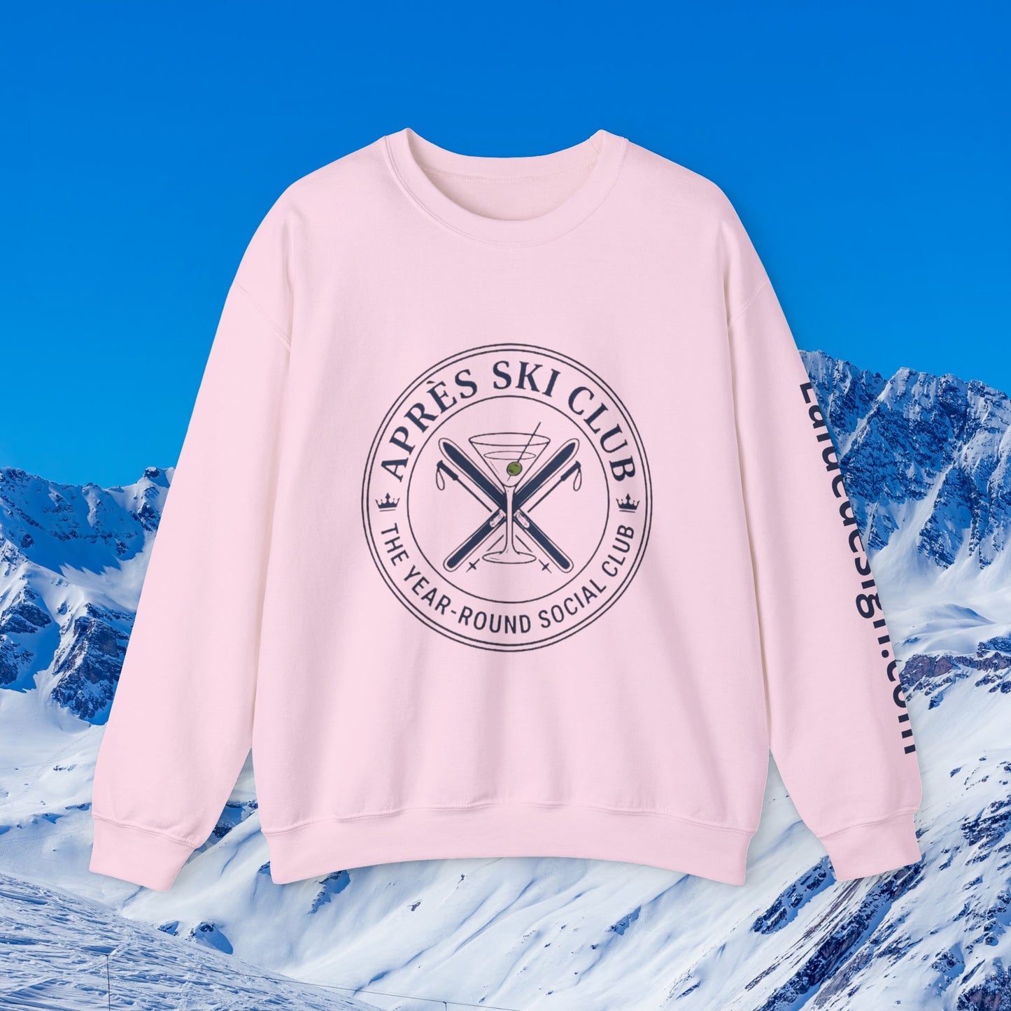 Après Ski Sweatshirt A light pink crewneck sweatshirt with a circular black graphic on the chest reading 'APRÈS SKI CLUB THE YEAR-ROUND SOCIAL CLUB' around crossed skis and a martini glass, plus black text down the right sleeve, shown against a snowy mountain and blue sky background.