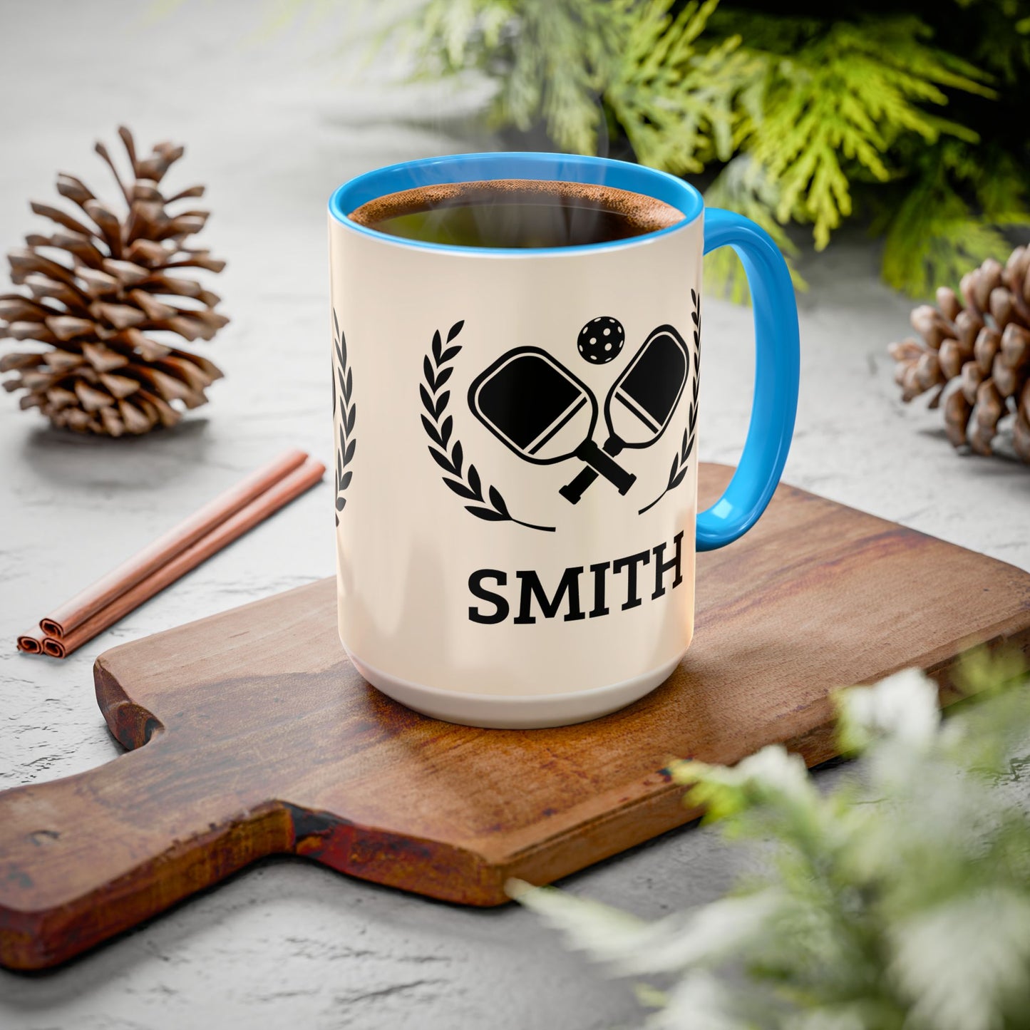 pickleball mug A ceramic mug with a blue rim and handle sits on a wooden cutting board. The mug is cream-colored with a black graphic of crossed ping-pong paddles and the name 'SMITH' printed below.