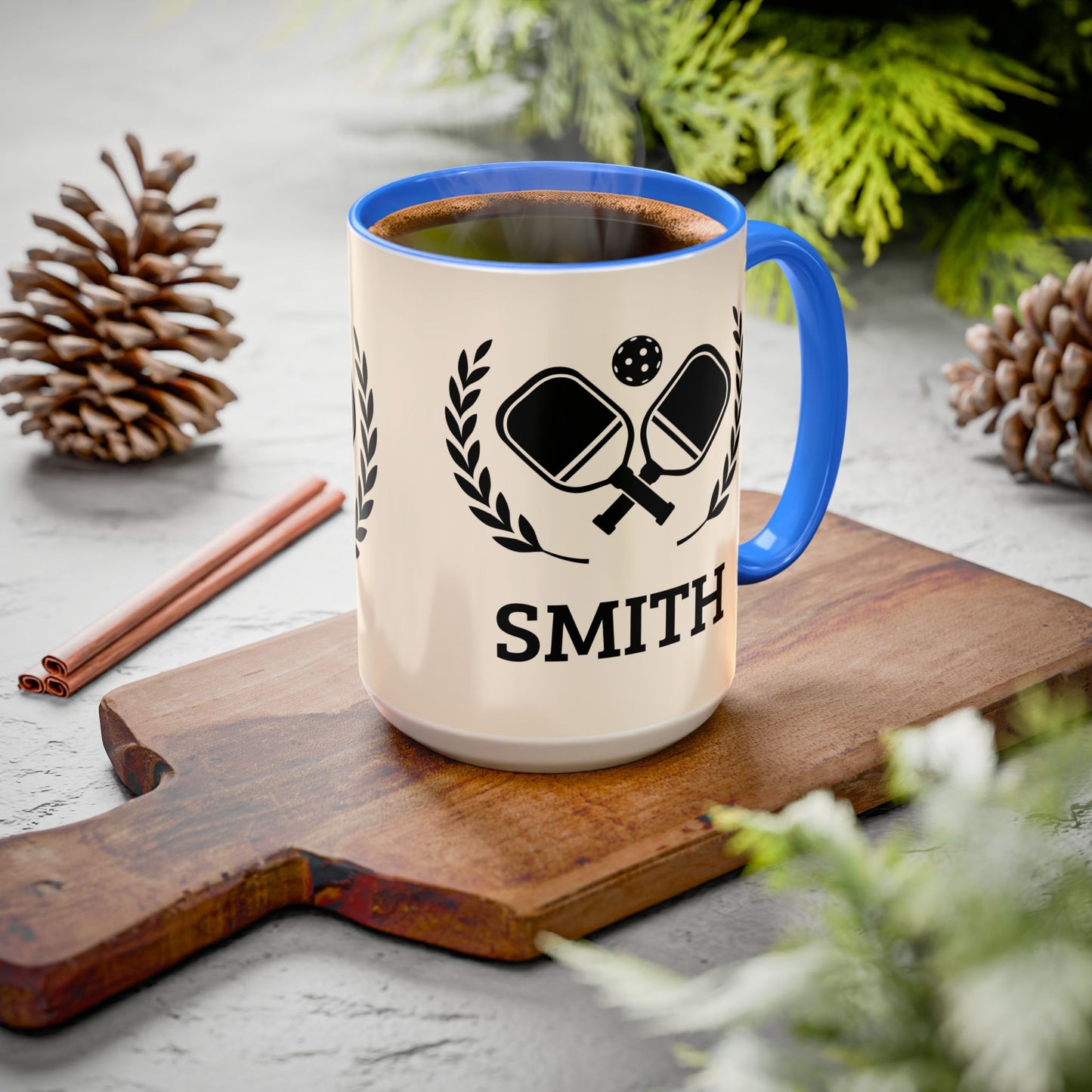 pickleball mug A ceramic mug with a blue rim and handle sits on a wooden cutting board. The mug is cream-colored with a black graphic of crossed ping-pong paddles and the name 'SMITH' printed below.