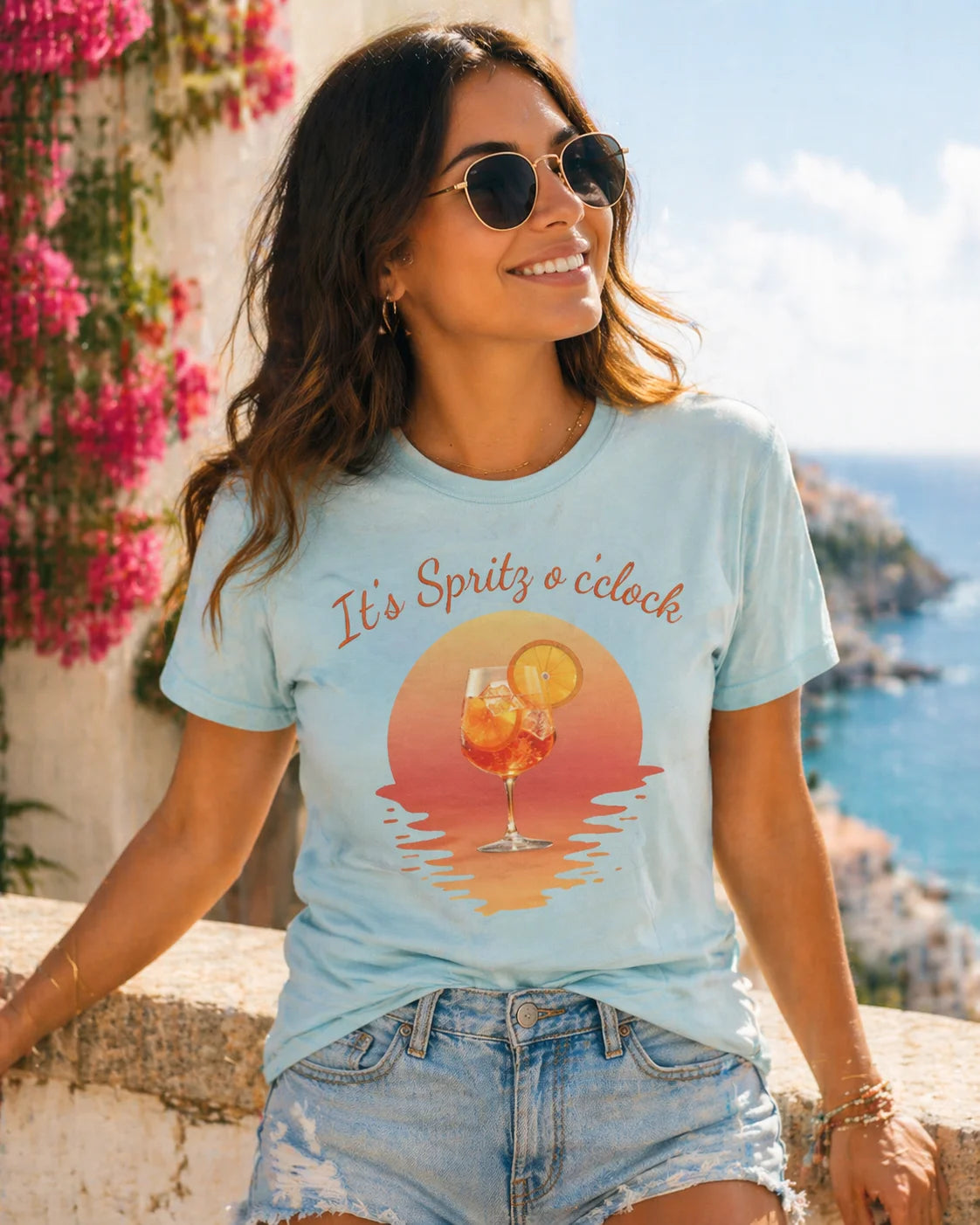Aperol T-Shirt Woman wearing a light blue t-shirt with a sunset graphic and text, sitting by a scenic location.