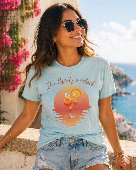Aperol T-Shirt Woman wearing a light blue t-shirt with a sunset graphic and text, sitting by a scenic location.