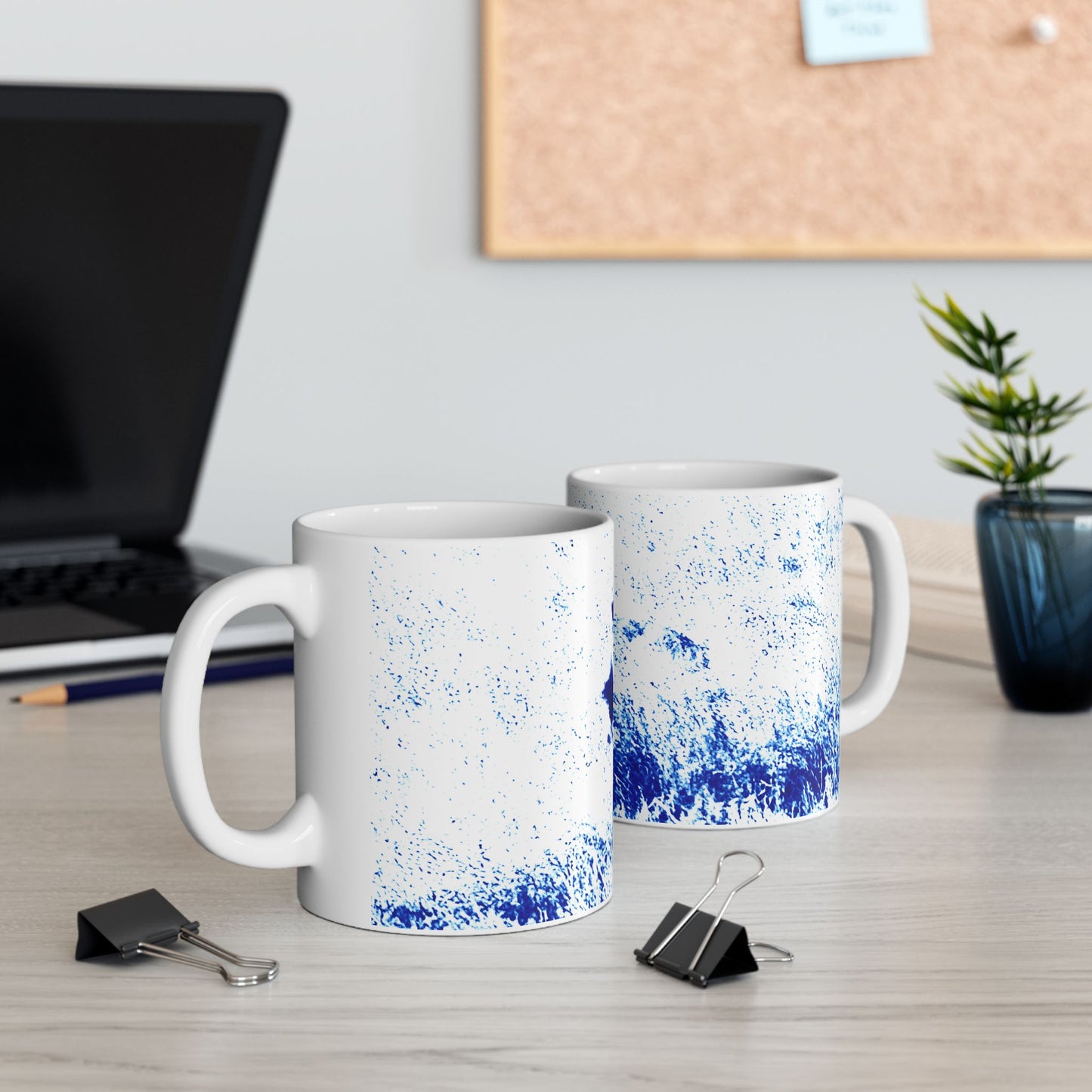 Two white mugs with blue speckles on a desk with office supplies and a plant.