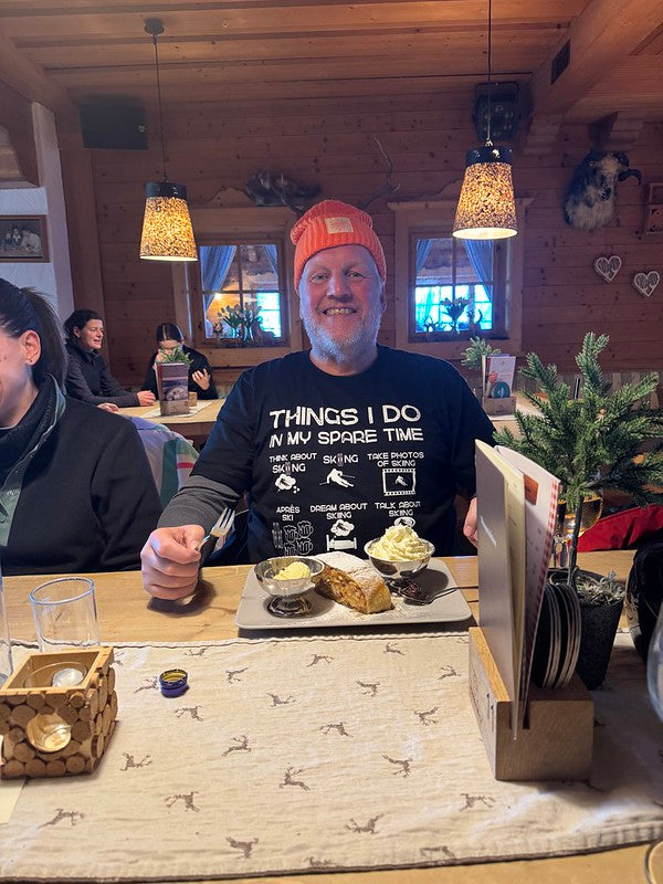 Laluedesign.com founder wearing the best-selling Ski T-shirt indoors in ski resort. Man sitting at a table in a cozy indoor setting with food and drinks.