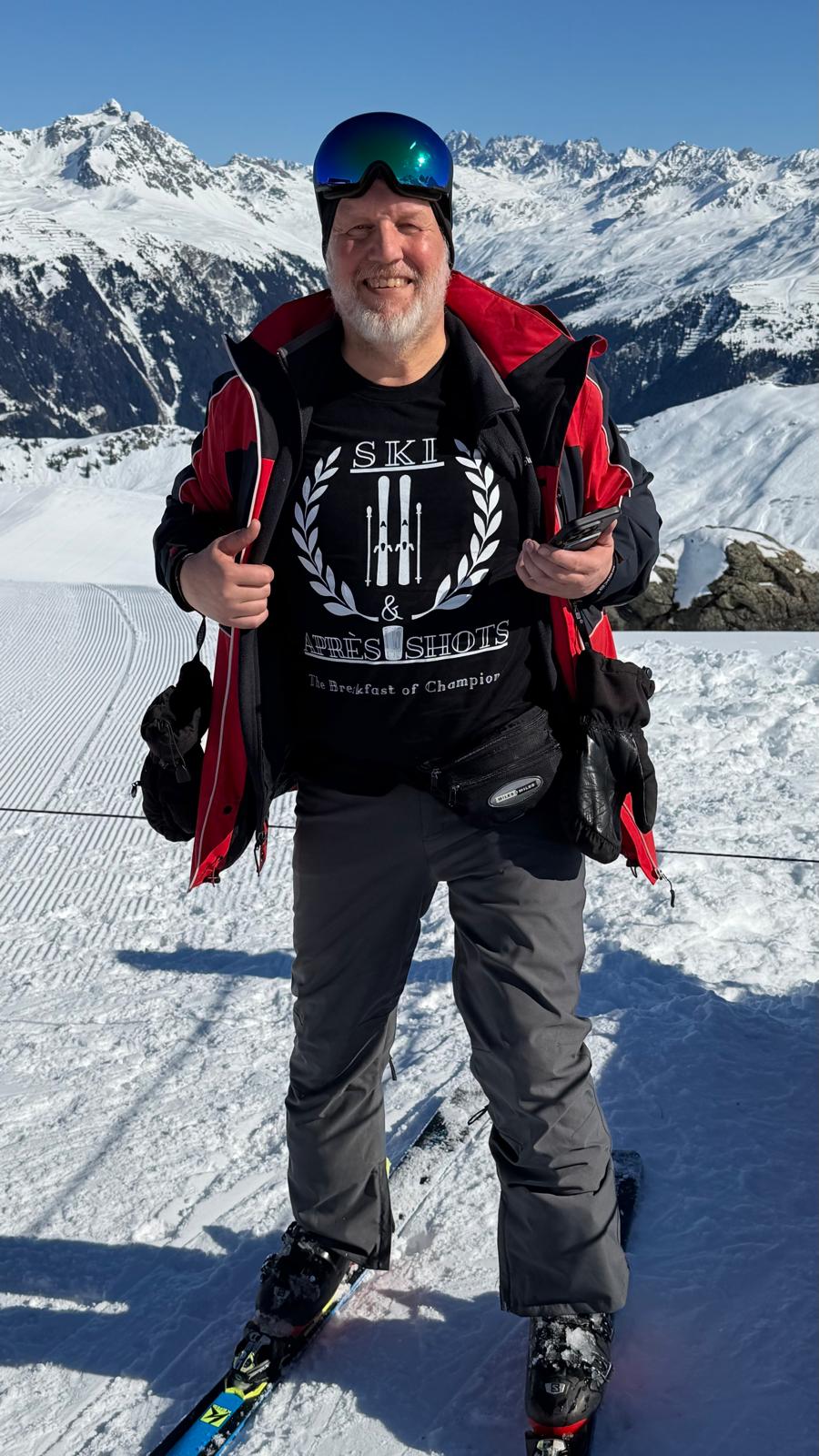 Laluedesign.com founder wearing the Ski and Apres shots T-shirt outdoors in ski resort putting the thumbs up.  in skiing gear with skis on a snowy mountain