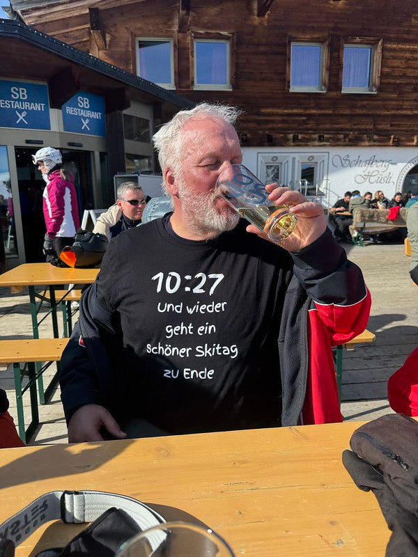 Laluedesign.com founder wearing the best-selling Ski T-shirt outdoors Man drinking from a glass at an outdoor restaurant with a wooden building in the background