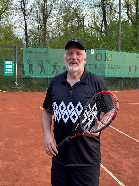 Laluedesign founder in diamond shape shirt with Tennis racket on a clay court with trees and blue sky in the background